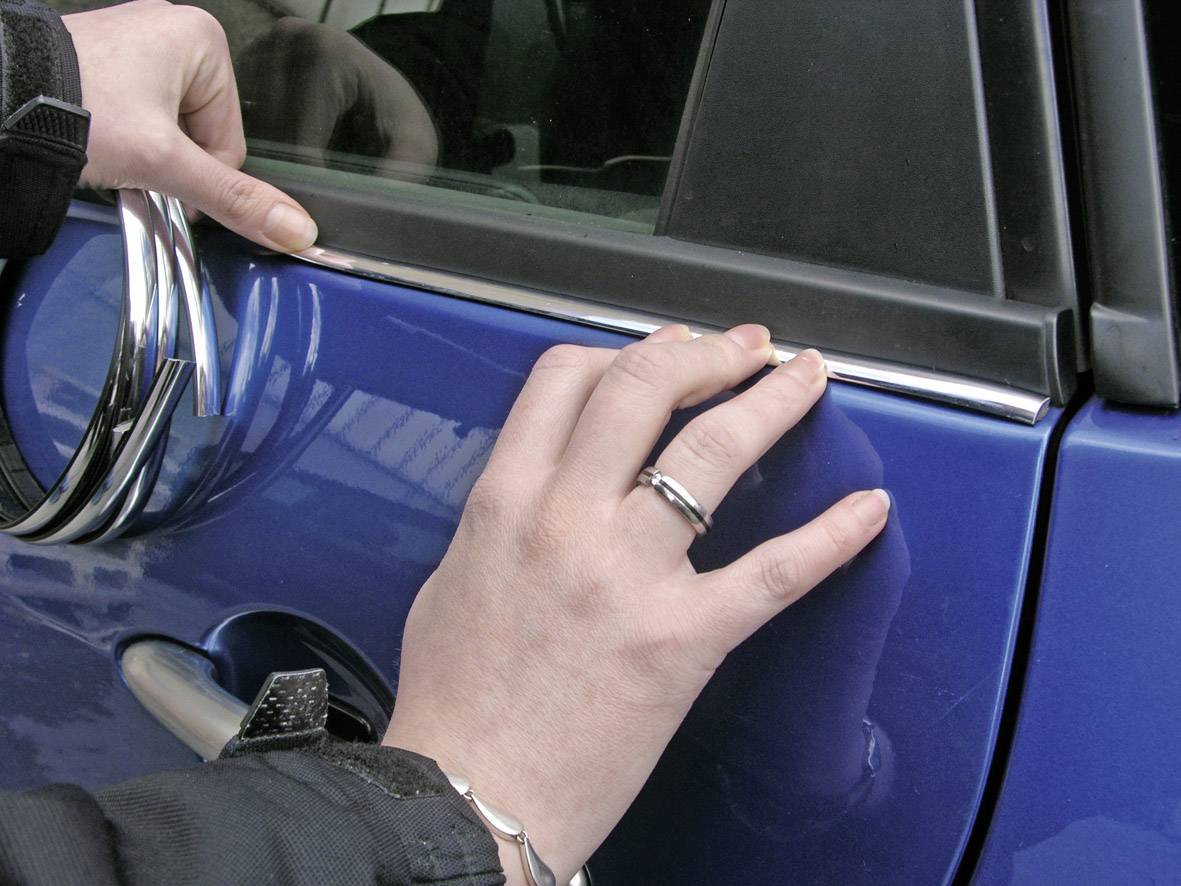 Iemand bevestigt een sierlijst aan de autodeur van een blauwe auto. Handen drukken de lijst stevig aan en richten deze uit.