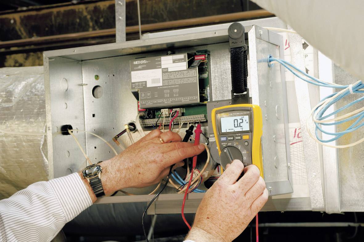 Een persoon meet de elektriciteit van een ventilatiesysteem met een geel multimeter die '0.2' aangeeft en controleert kabelverbindingen.
