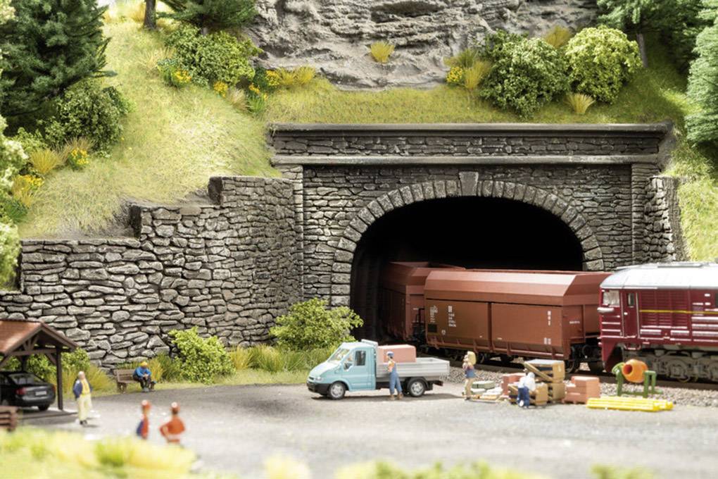 Een modeltreintje rijdt uit een stenen tunnel in een landschap met bomen en figuren die dicht bij een kleine, blauwe vrachtwagen staan.