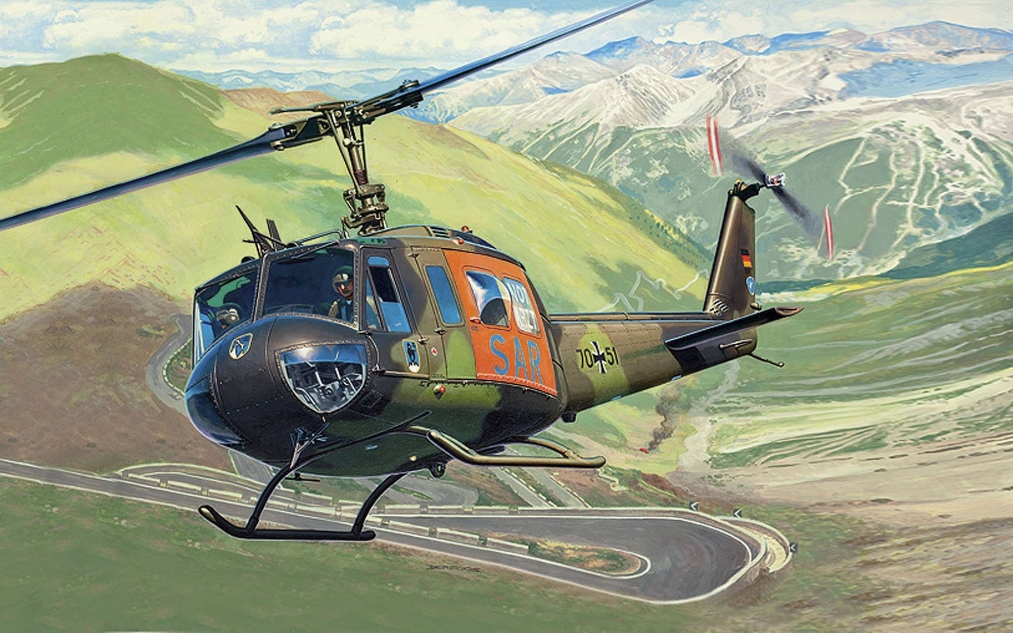 Een helikopter vliegt over een bergachtig landschap met een SAR-logo aan de zijkant. Op de achtergrond zijn slingerende wegen en met sneeuw bedekte bergen te zien.