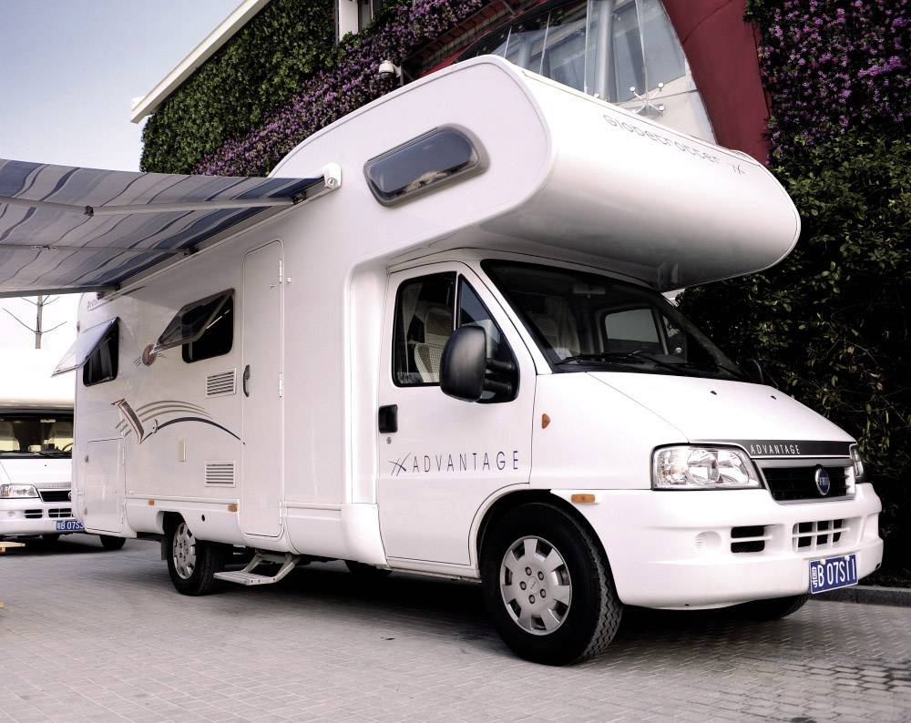 Camper staat geparkeerd op een bestrate parkeerplaats met uitgeschoven luifel. Op de achtergrond is een modern gebouw zichtbaar.
