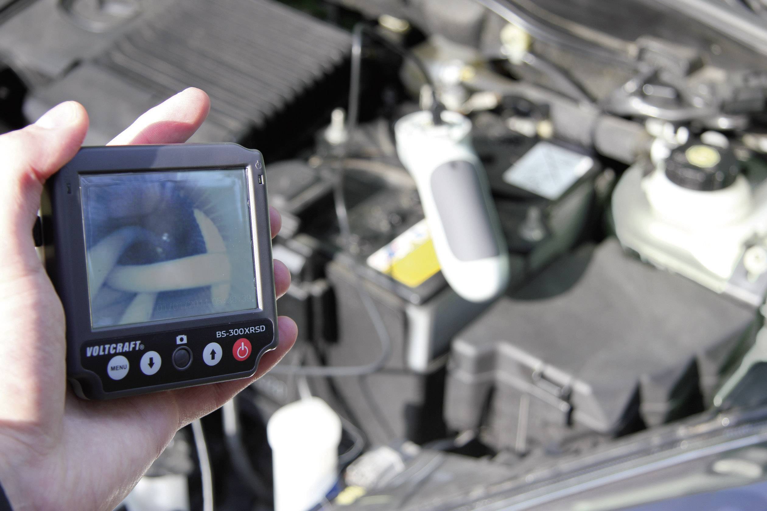 Een persoon houdt een diagnoseapparaat met een scherm vast, dat de interne toestand van een auto-onderdeel weergeeft. Op de achtergrond is de motorruimte zichtbaar.