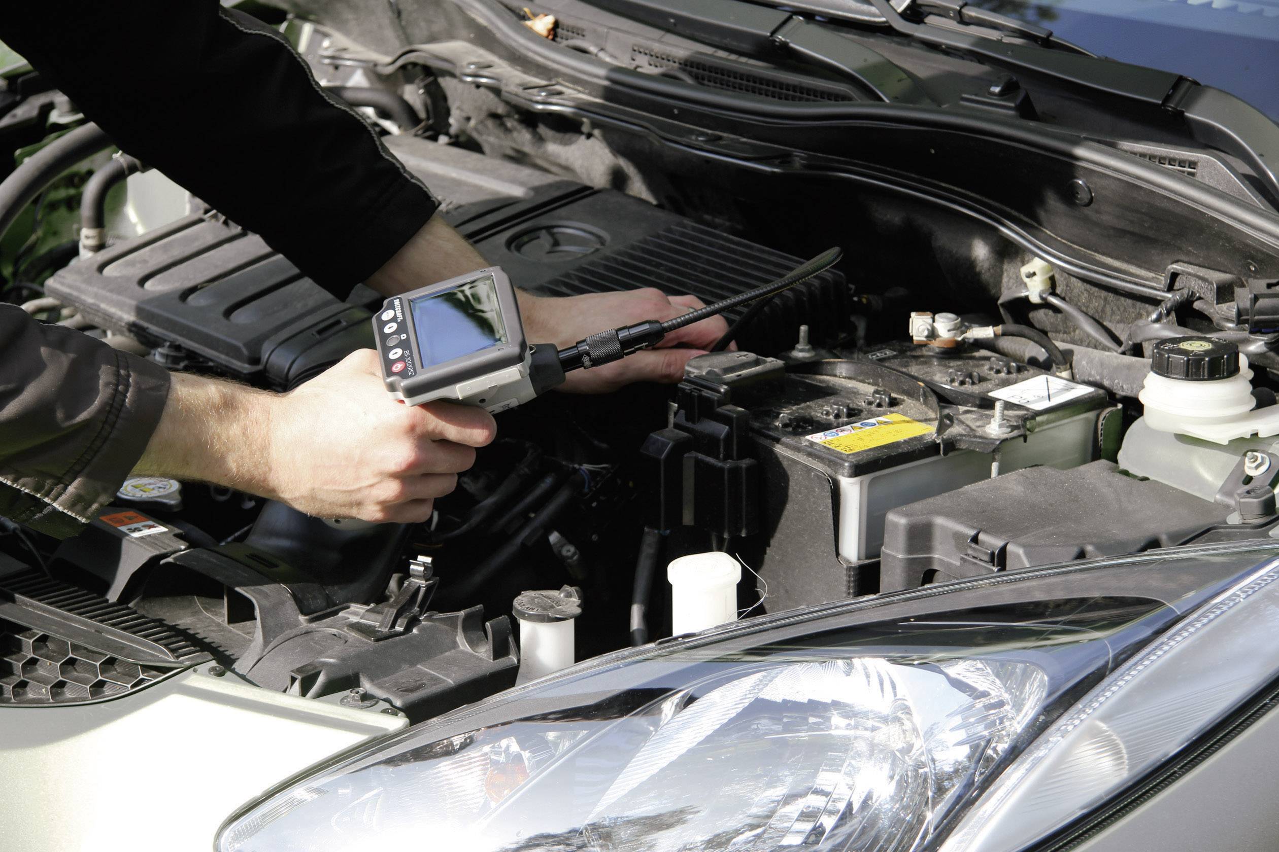 Een persoon controleert de motor van een auto met een diagnoseapparaat. De motorkap staat open, en het apparaat toont gegevens op een scherm.