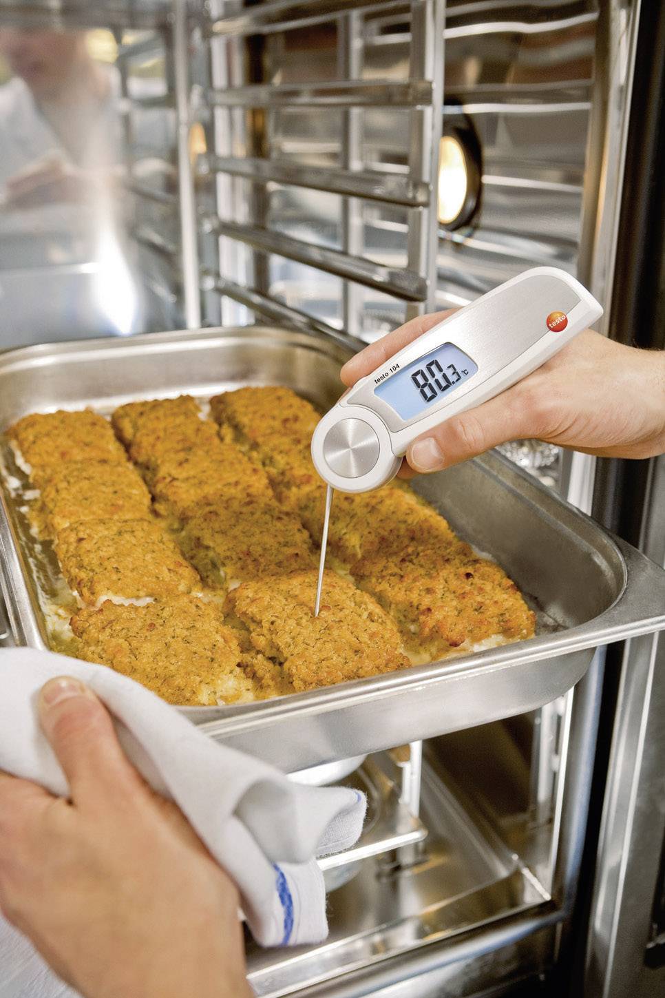 Een persoon meet de kerntemperatuur van gehaktbroodjes op een bakplaat in de oven met een digitaal thermometer die 80,3°C aangeeft.