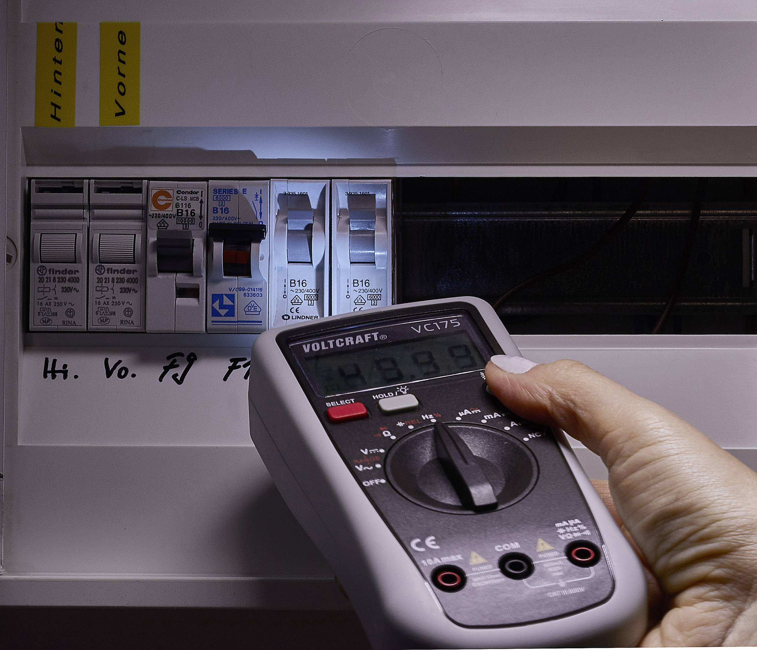 Een hand houdt een digitale multimeter voor een open zekeringkast om de spanning te meten. De display toont '234.9'.