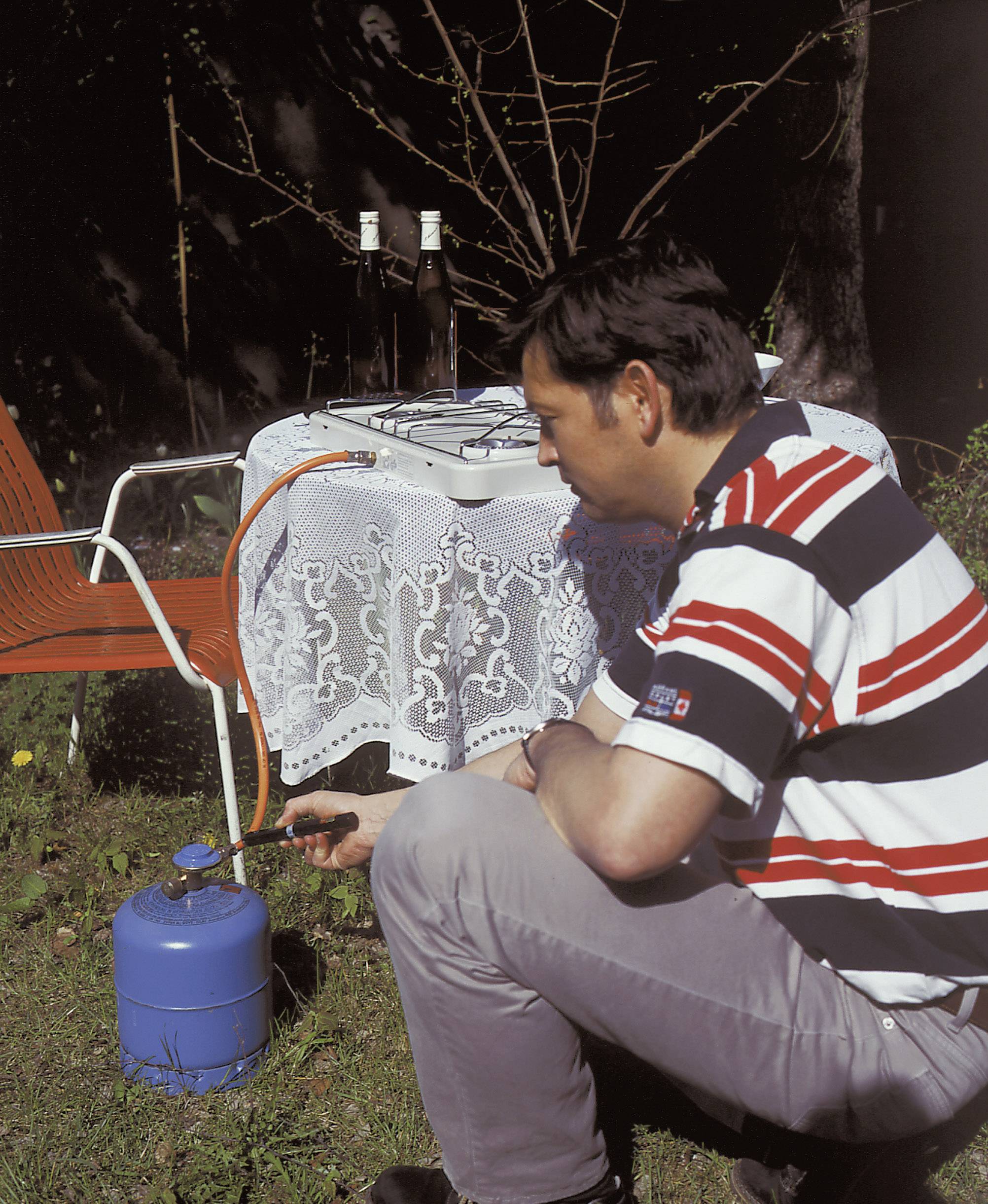 Een man steekt een blauwe gasstel aan met een aansteker in de buitenlucht. Op de achtergrond staat een tafel met een tafelkleed en twee flessen.