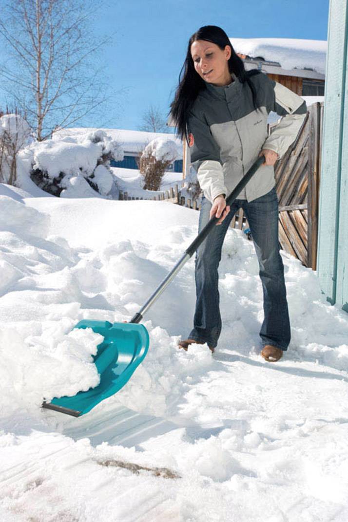 Een vrouw schept sneeuw voor een huis op een zonnige winterdag, met een blauwe lucht op de achtergrond.