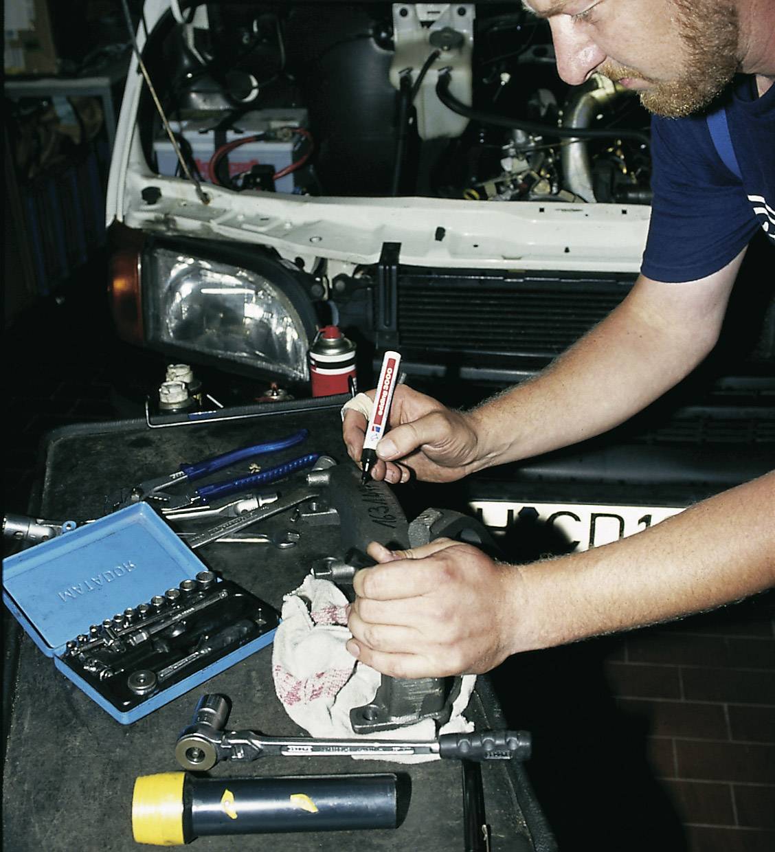 Een man werkt aan een auto en controleert een onderdeel. Op tafel liggen verschillende gereedschappen en een open gereedschapskist.