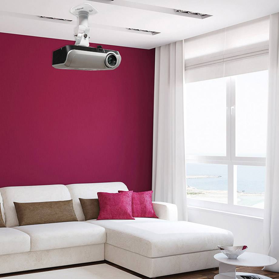 Woonkamer met een witte bank, rode kussens en een projector aan het plafond. Een groot raam biedt uitzicht op zee.