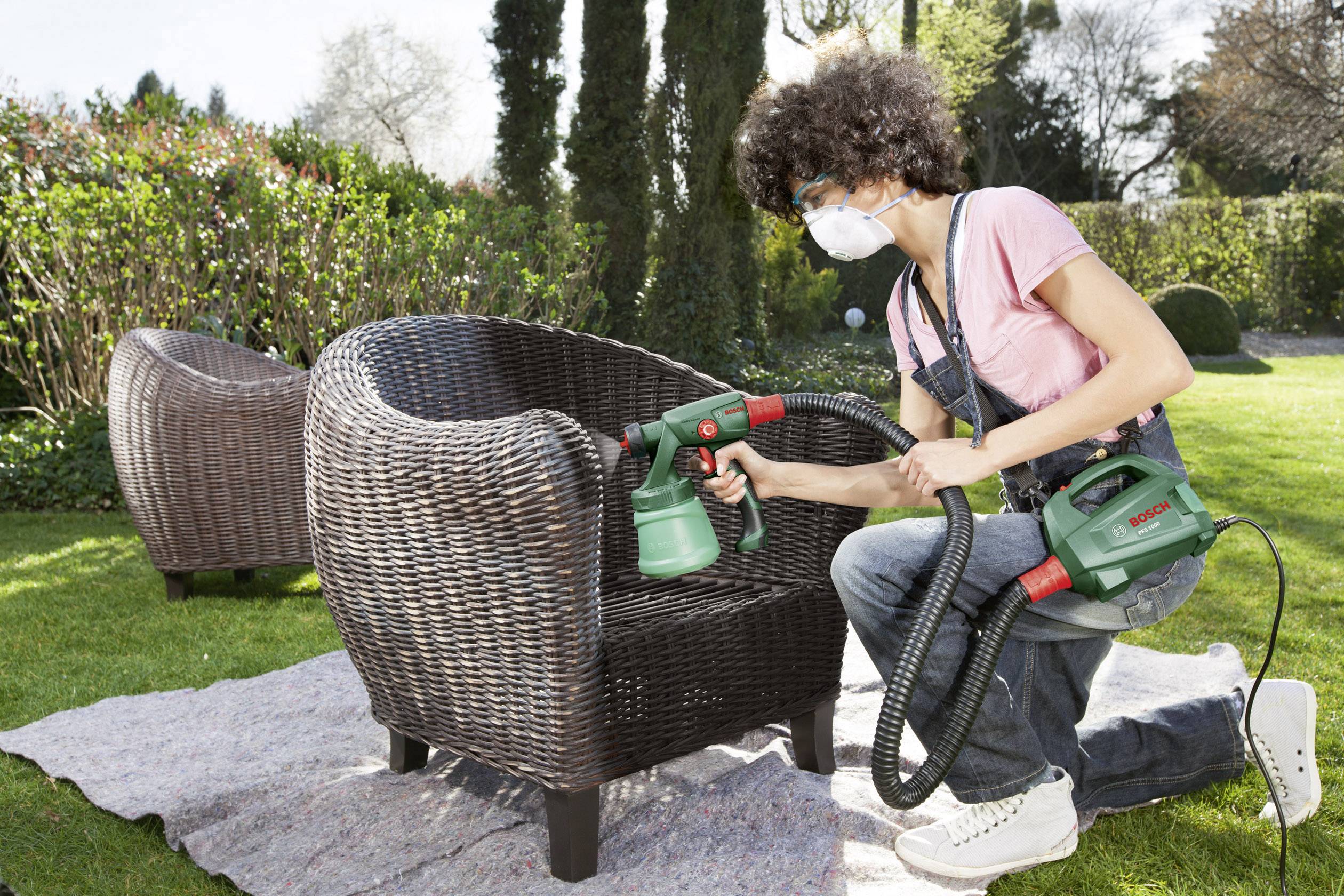 Een persoon spuit verf op een tuinstoel, draagt daarbij een veiligheidsbril en een masker. Op de achtergrond bevindt zich een tuin.