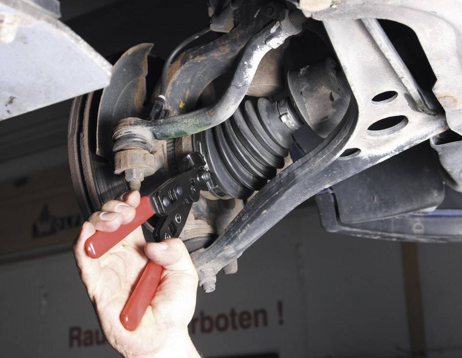 Een hand gebruikt een tang met rode handgrepen om onderdelen van een voertuigonderstel te repareren. Op de achtergrond zijn onderdelen van een werkplaats zichtbaar.