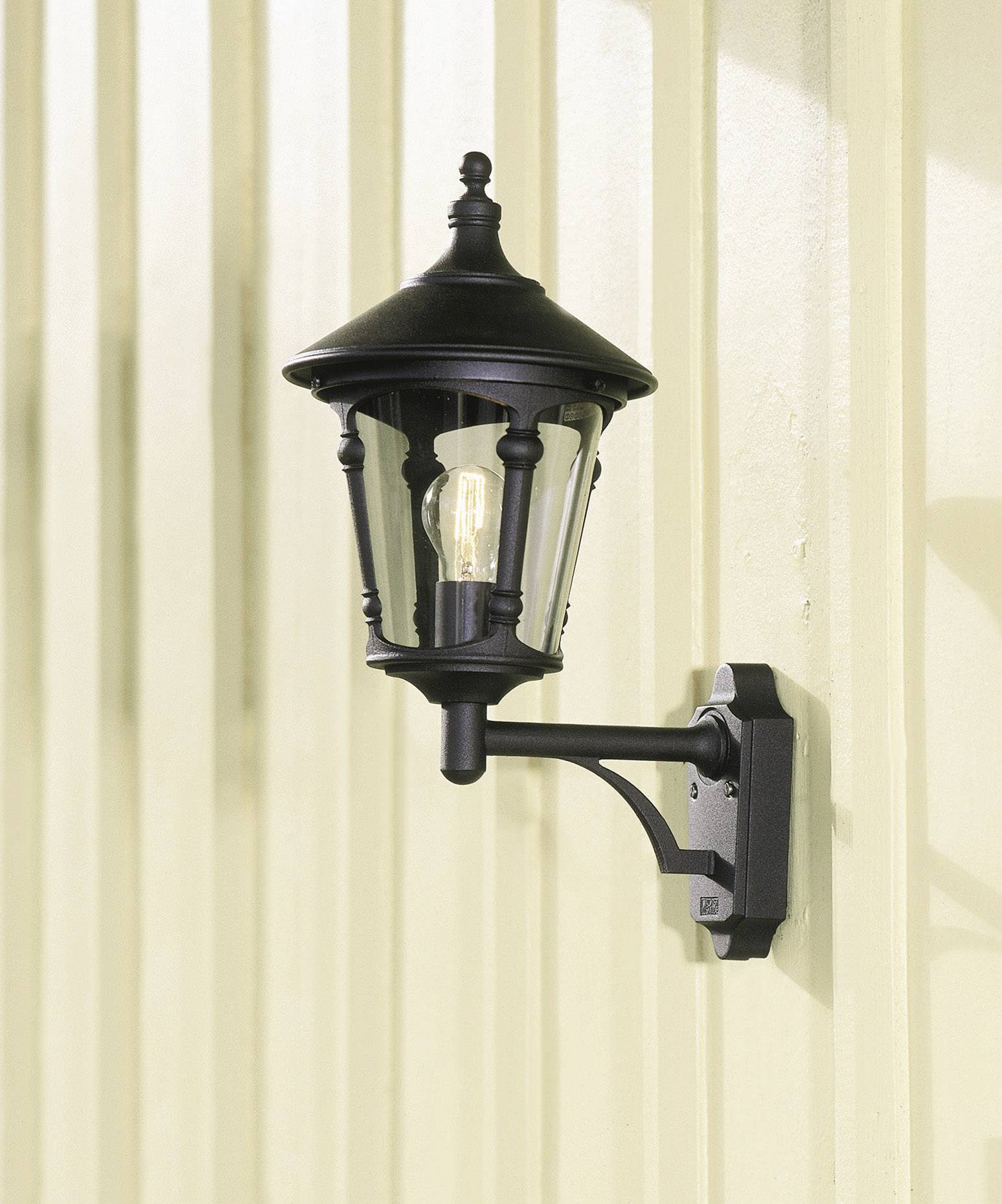 Zwarte buitenlantaarn gemonteerd aan een lichtgele muur, met een glazen kap waarachter de gloeilamp zichtbaar is.