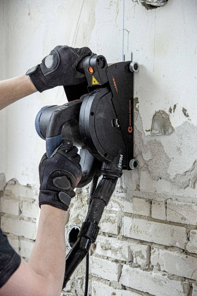 Een persoon gebruikt een muurfreesmachine om sleuven in een bakstenen muur te snijden. Beschermende kleding zoals handschoenen wordt gedragen.