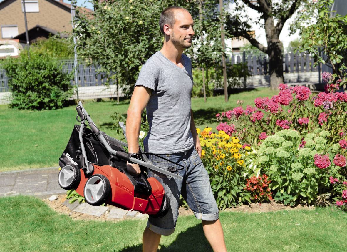 Een man draagt een opgevouwen grasmaaier door een bloeiende tuin. Het is een zonnige dag.
