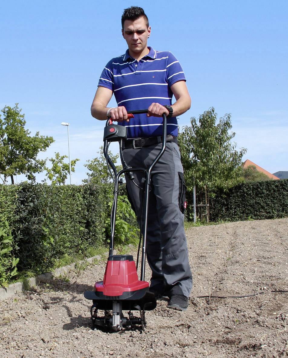 Een man in een blauw gestreept poloshirt bedient een rode tuinfrees op een veld. Op de achtergrond zijn bomen en een blauwe lucht.