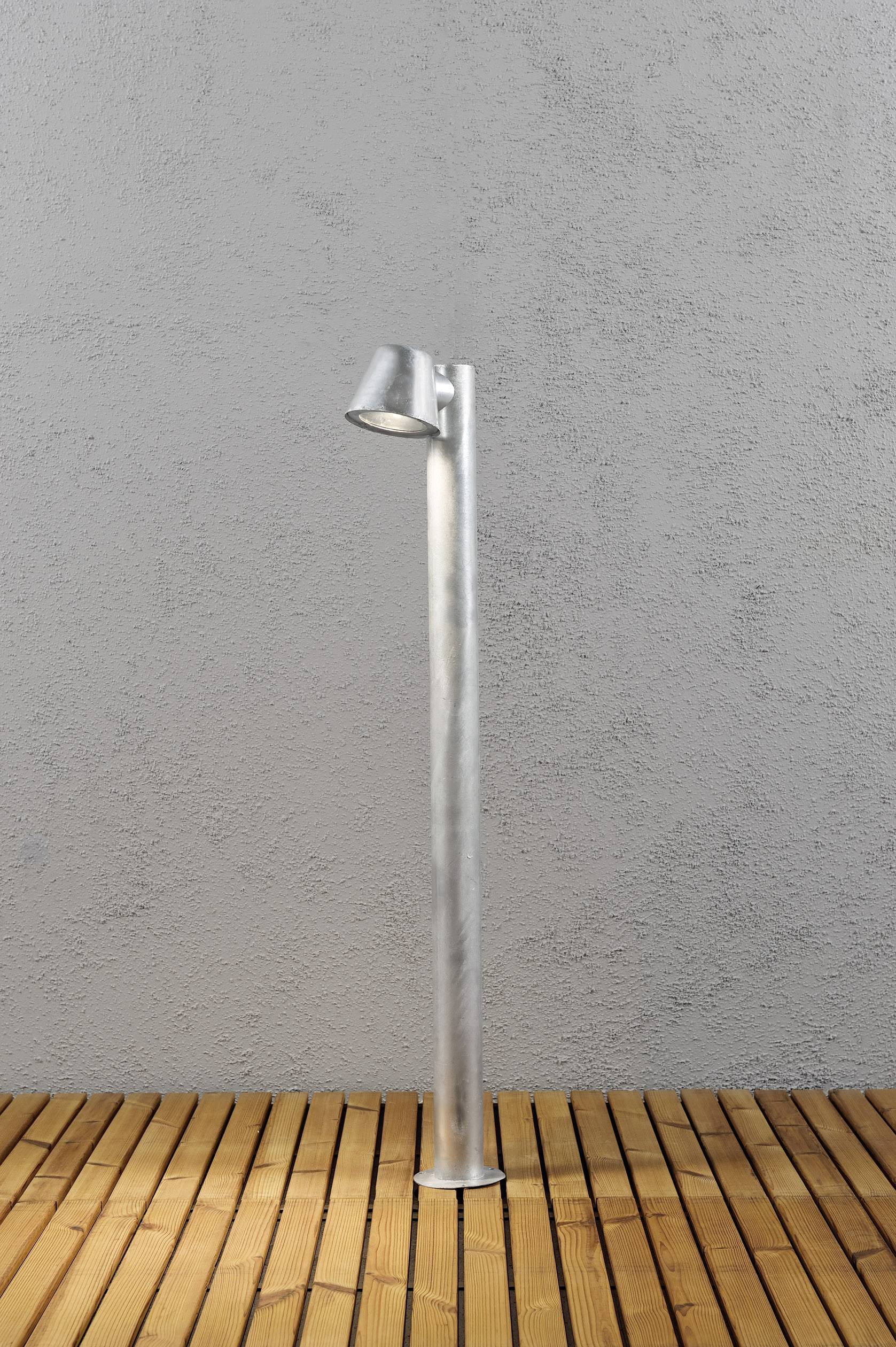 Een zilveren buitenlamp op een houten terras voor een grijze muur. De lamp is rond en heeft een smal, hoog voetstuk.