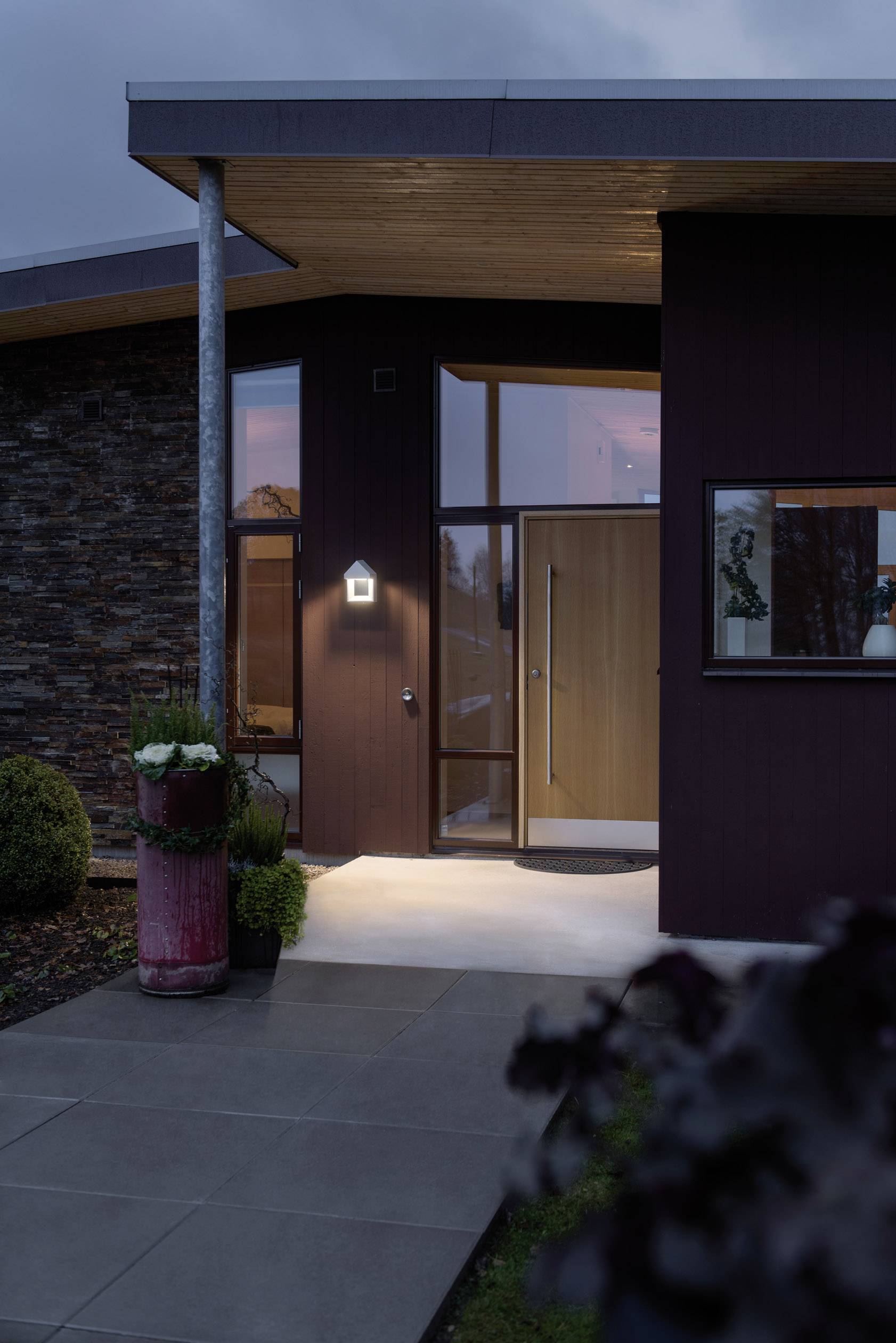 Modern huis met donkere gevel en grote ramen, verlicht door een buitenlamp, voor een bewolkte avondlucht.