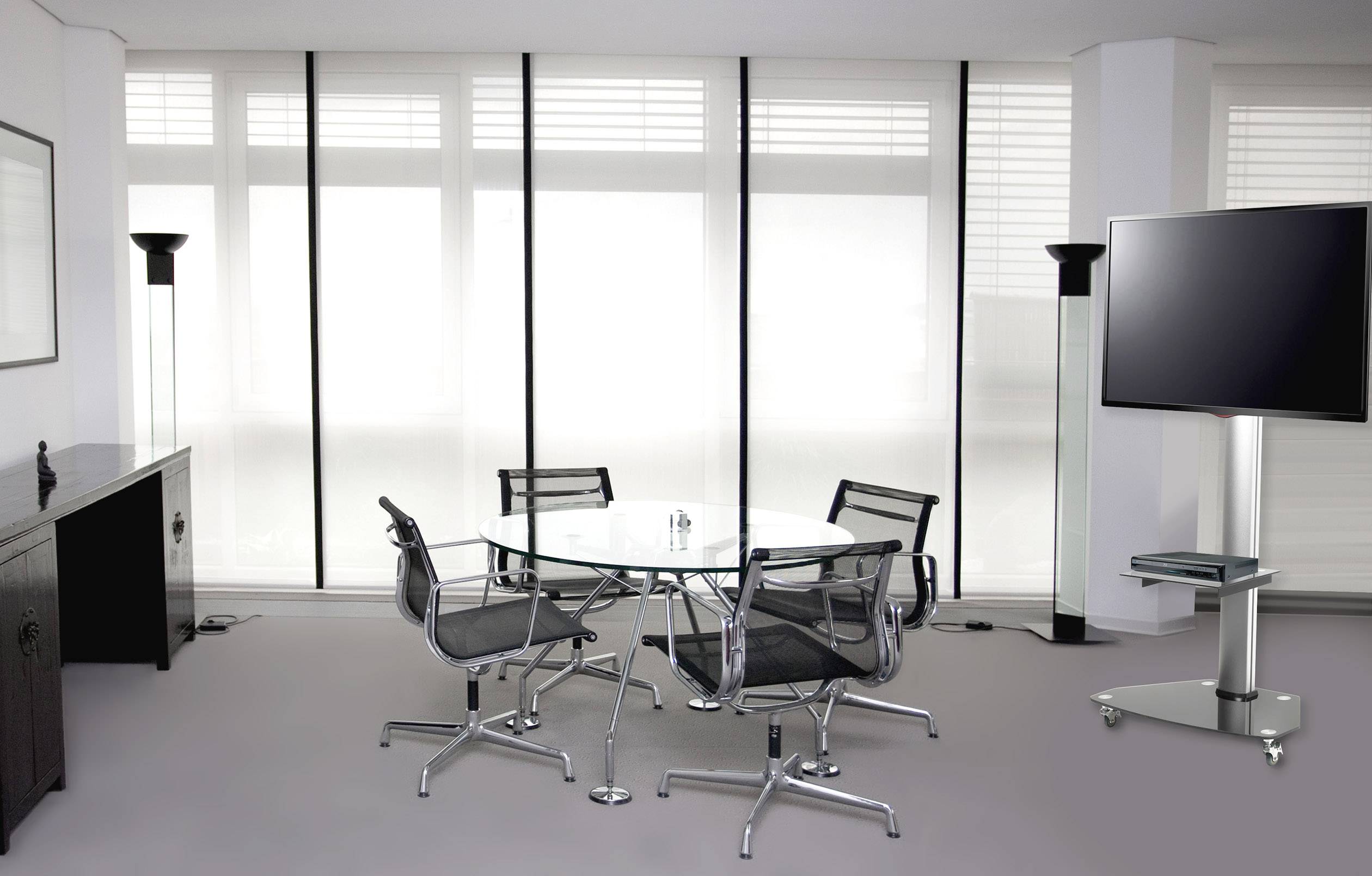 Een moderne vergaderruimte met een ronde glazen tafel en vier zwarte stoelen, een groot flatscreen-scherm op een standaard.