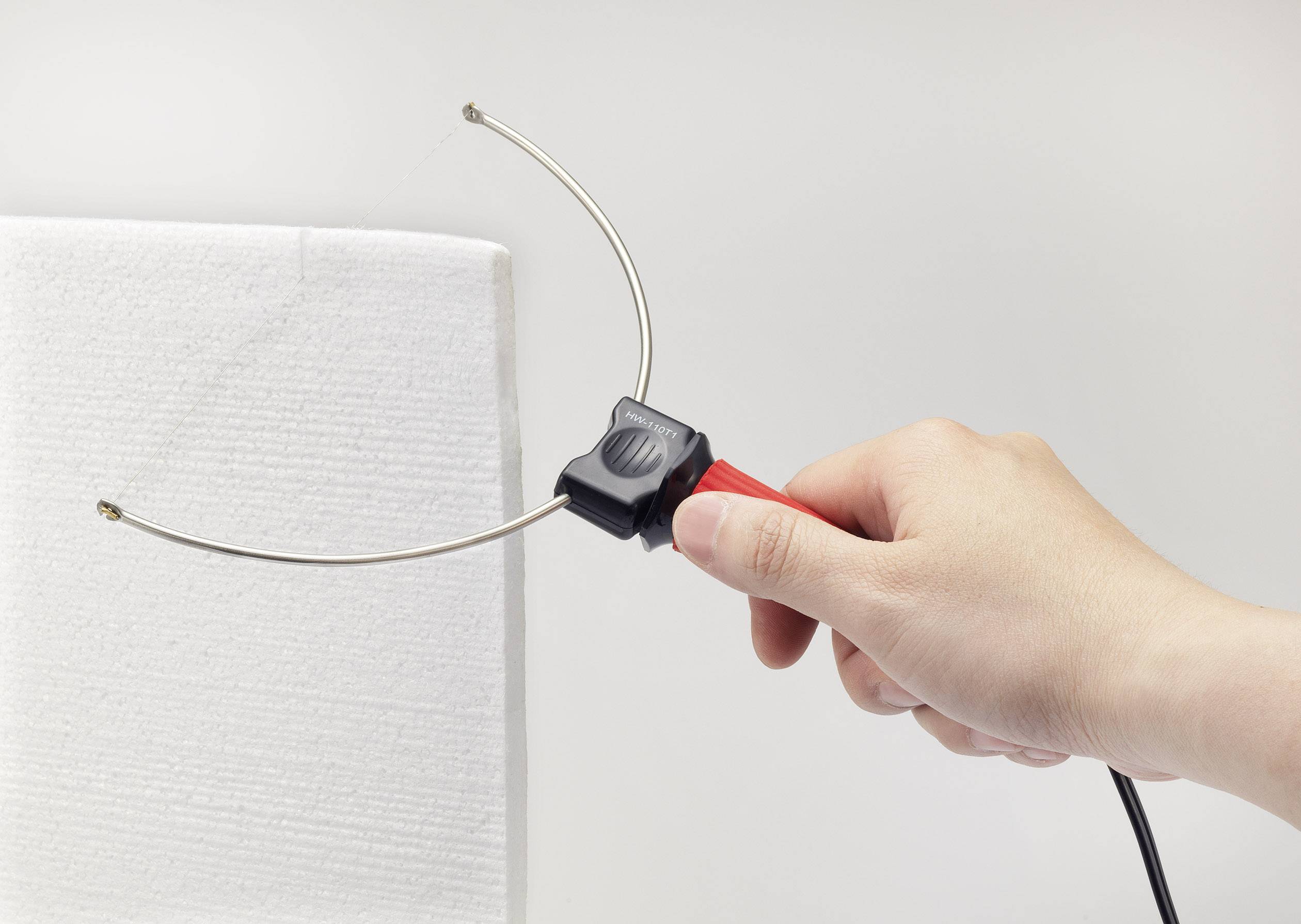 Een hand houdt een heet snijgereedschap vast dat een piepschuimplaat doorsnijdt. Het gereedschap heeft twee gebogen metalen stangen.