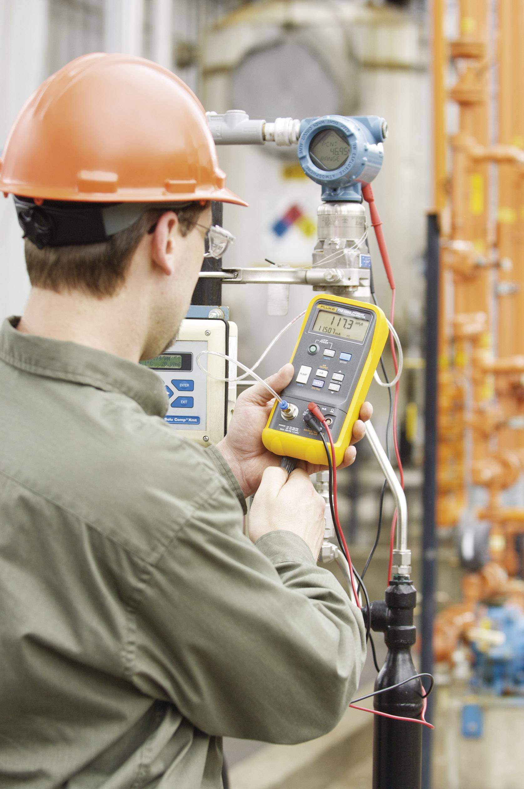 Een man met een veiligheidshelm controleert technische apparatuur met een multimeter. Hij staat in een industriële omgeving met buizen op de achtergrond.