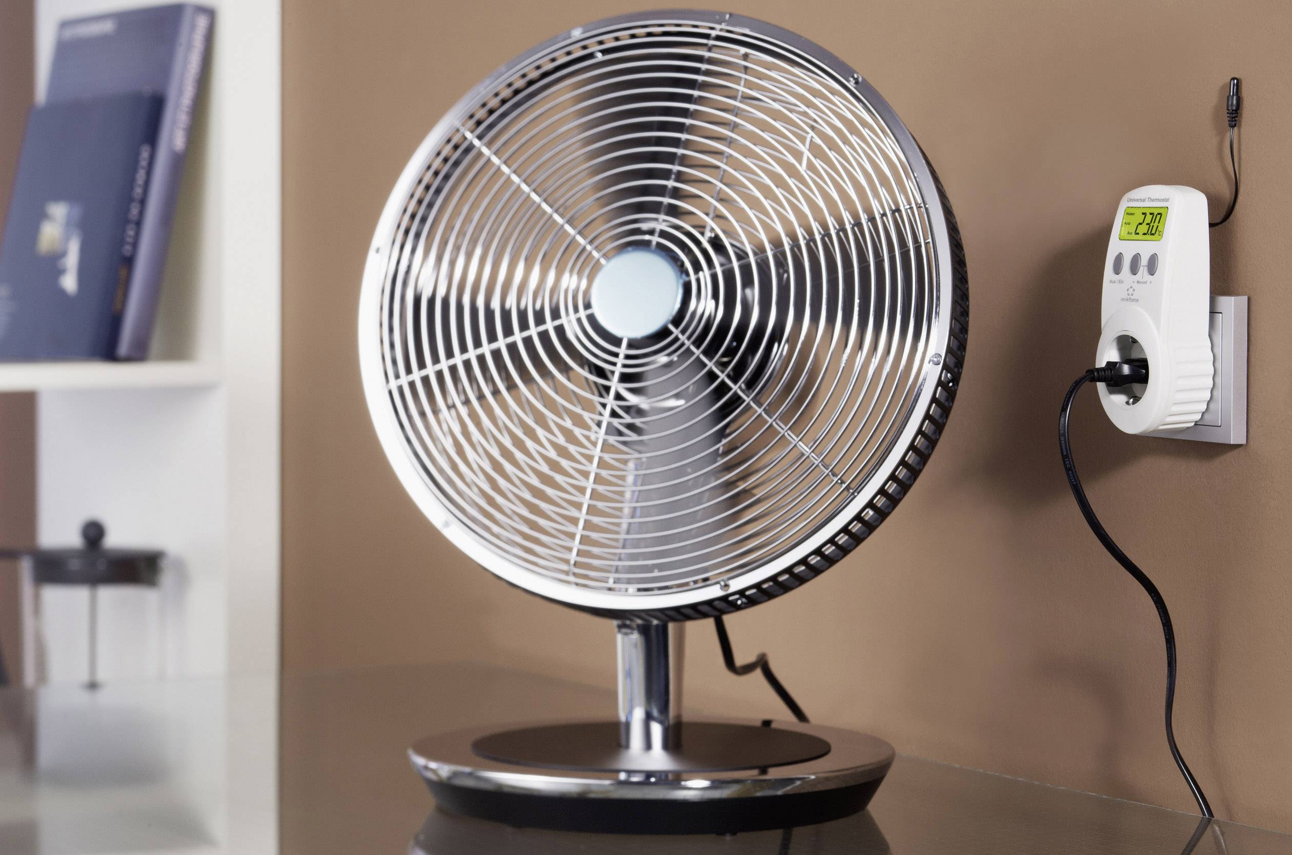 Een tafelventilator staat op een tafel naast een stopcontact, waaraan een digitaal stroommetingsapparaat is aangesloten.