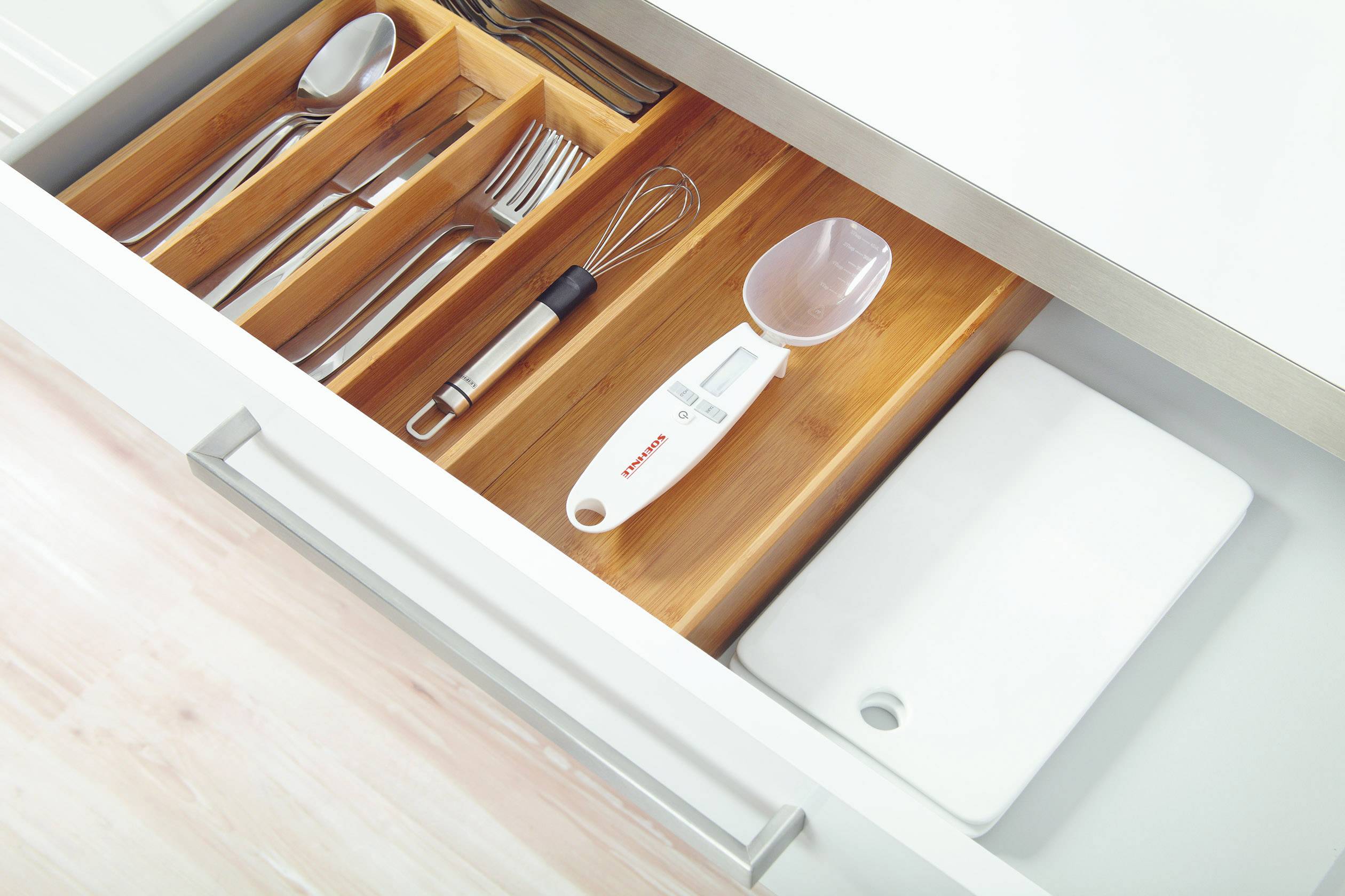 Openstaande keukenlades met houten vakken waarin bestek, een garde, een thermometer en een snijplank netjes zijn opgeborgen.