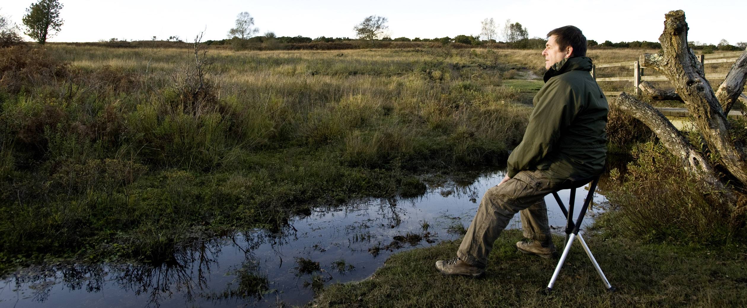 Een persoon zit op een opvouwbare stoel en kijkt uit over een rustig, natuurlijk landschap met water en bomen op de achtergrond.