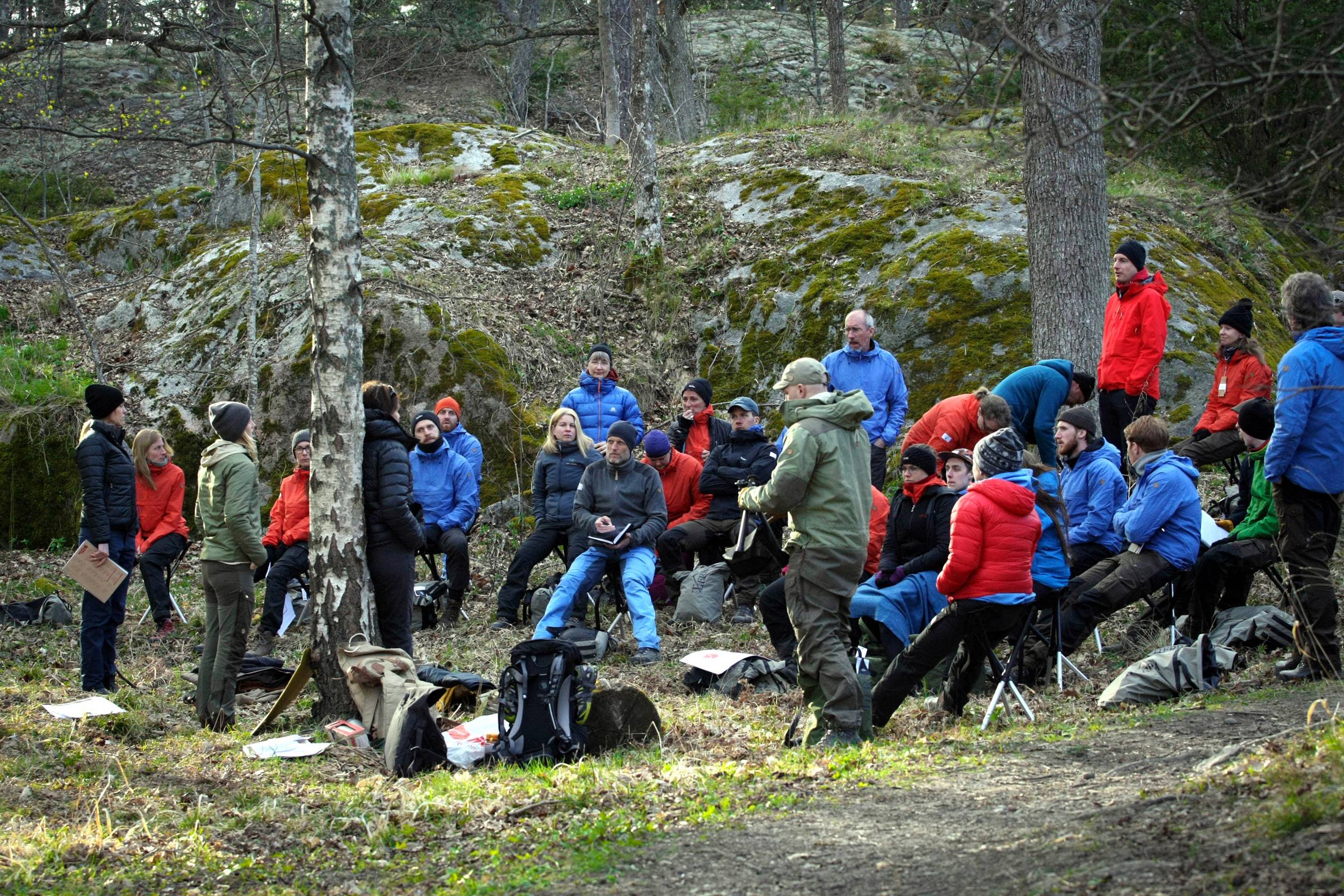 Bijeenkomst van mensen in een bos tijdens een vergadering of training. Enkele deelnemers dragen kleurrijke outdoorkleding, omringd door bomen en rotsen.