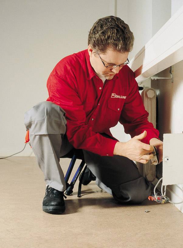 Een technicus in een rood shirt en grijze broek werkt aan een elektrische doos aan de muur.