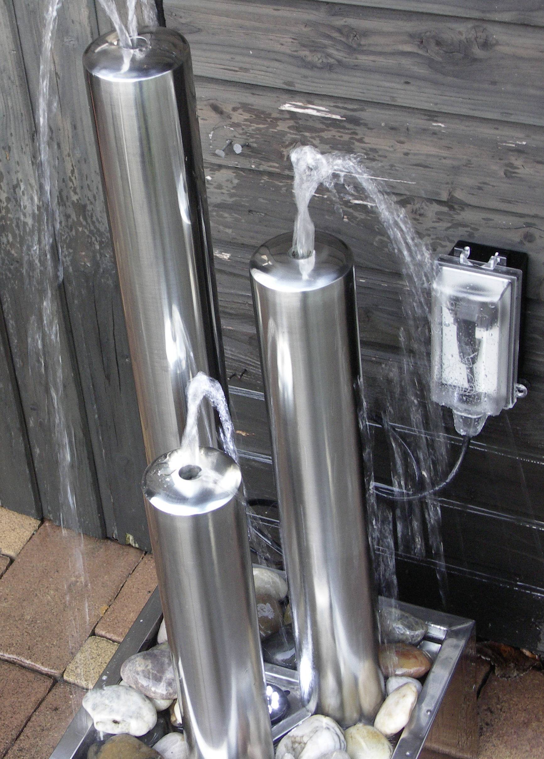 Drie zilveren waterfontijnen van verschillende hoogtes spuiten water in een tuingedeelte, omgeven door stenen.