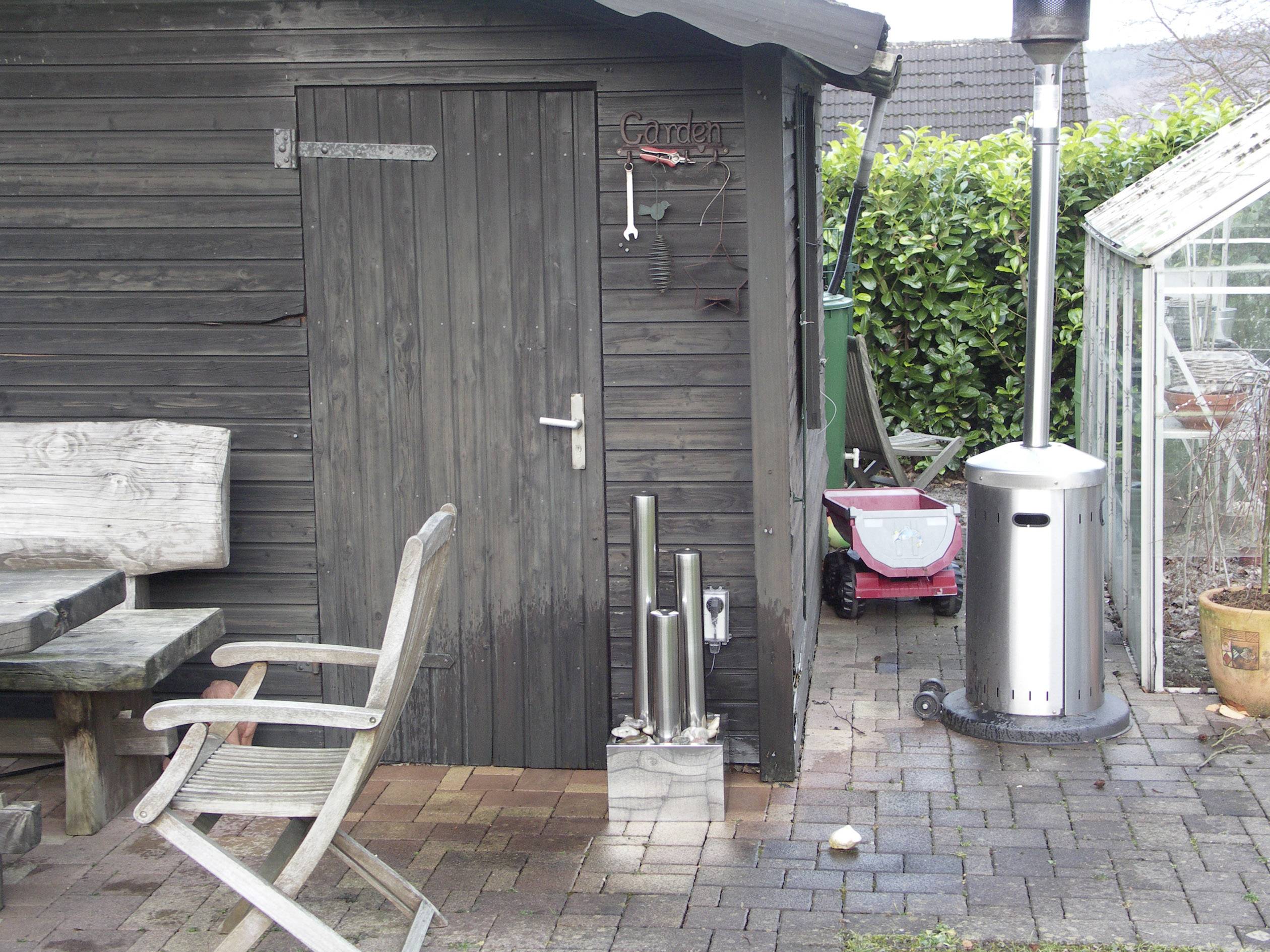 Houten schuur met bank, tafel en haard. Op de achtergrond een tuinhakselaar, een oven en een kas. Landelijk tuintafereel.