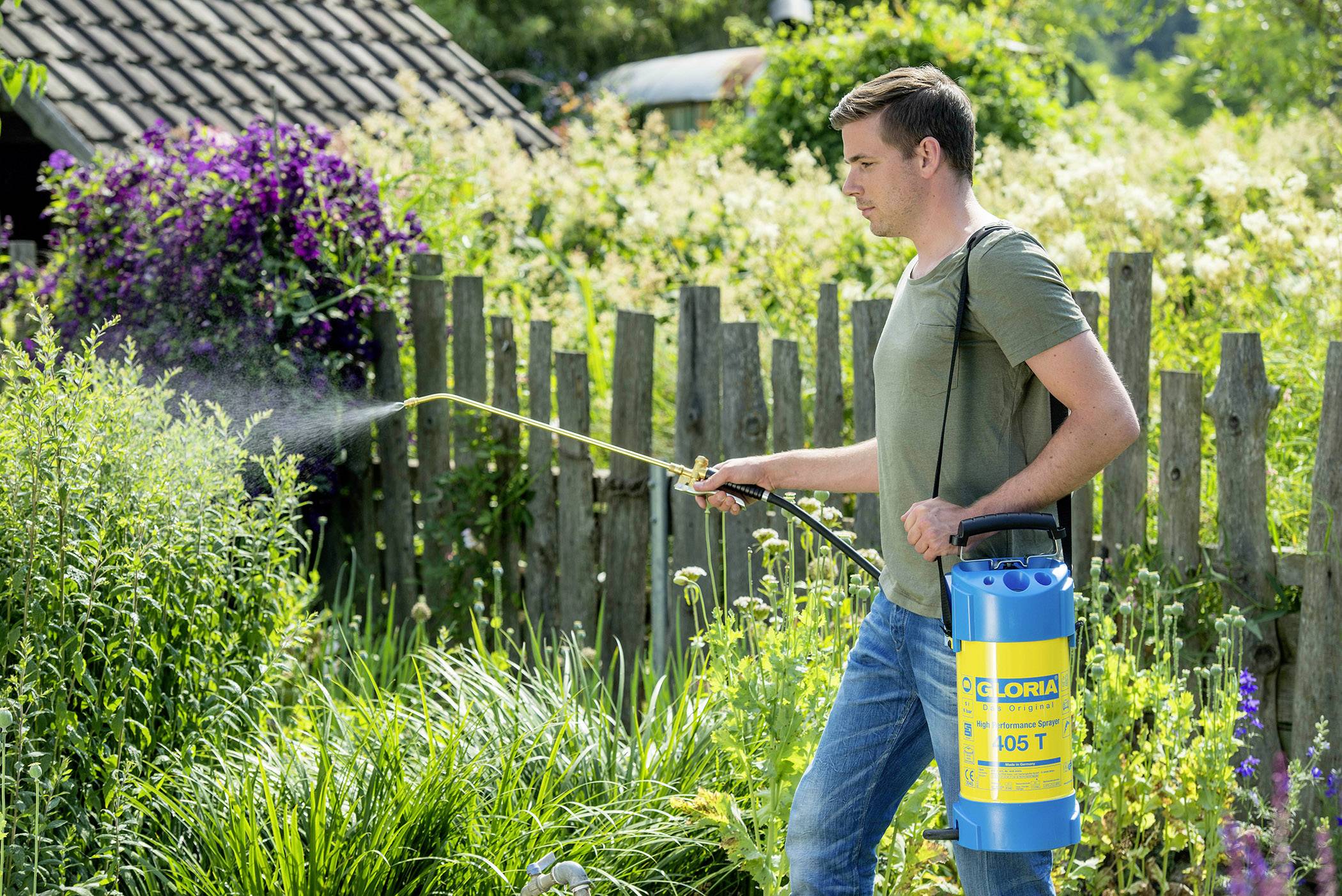 Man in groen T-shirt besproeit planten in de tuin met een blauwe drukspuit. Op de achtergrond is een houten hek en groene vegetatie te zien.