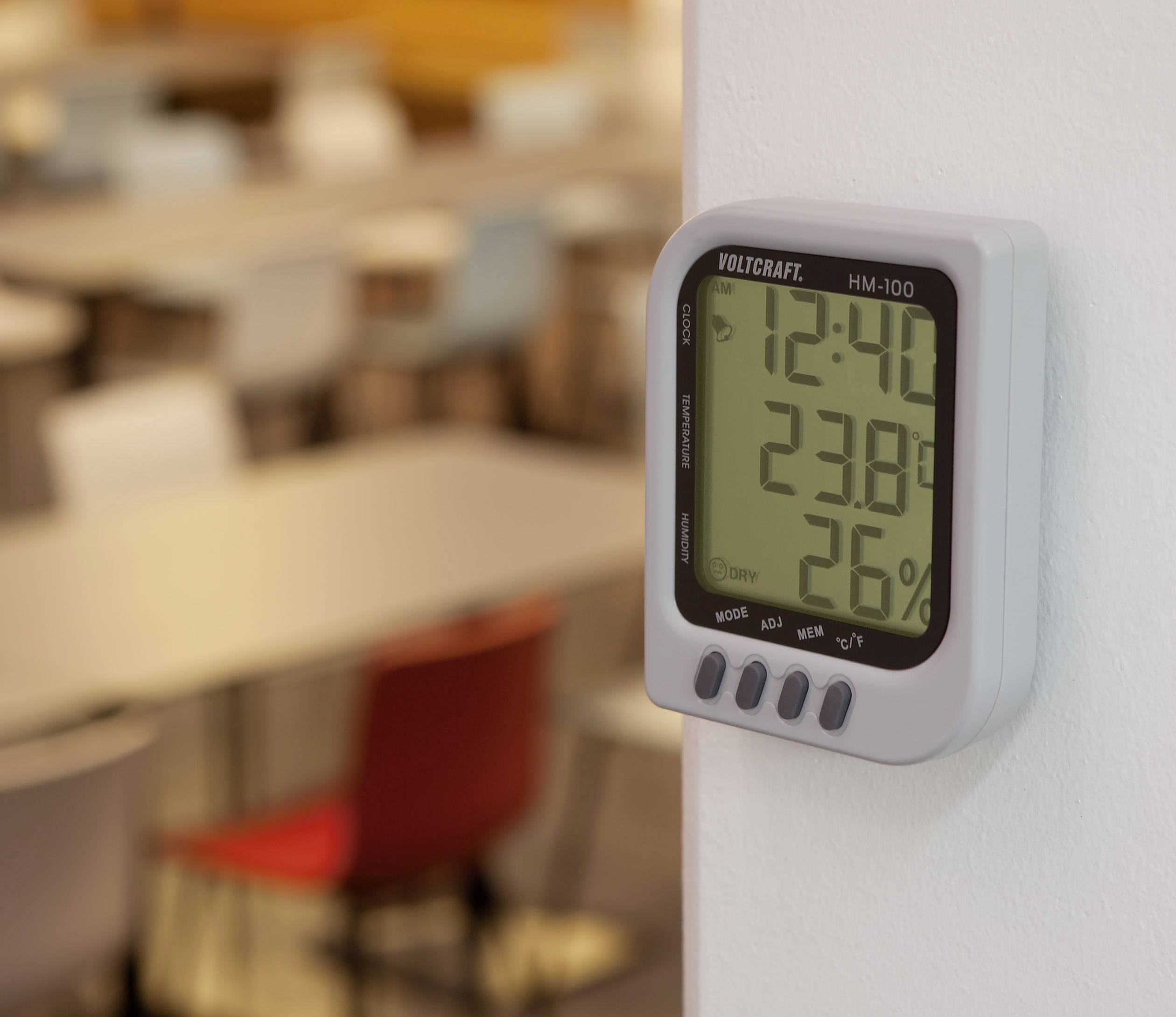 Een digitale thermo-hygrometer toont 12:40 uur, 23,8 graden Celsius en 26% luchtvochtigheid. Op de achtergrond zijn wazig bureau's en stoelen.