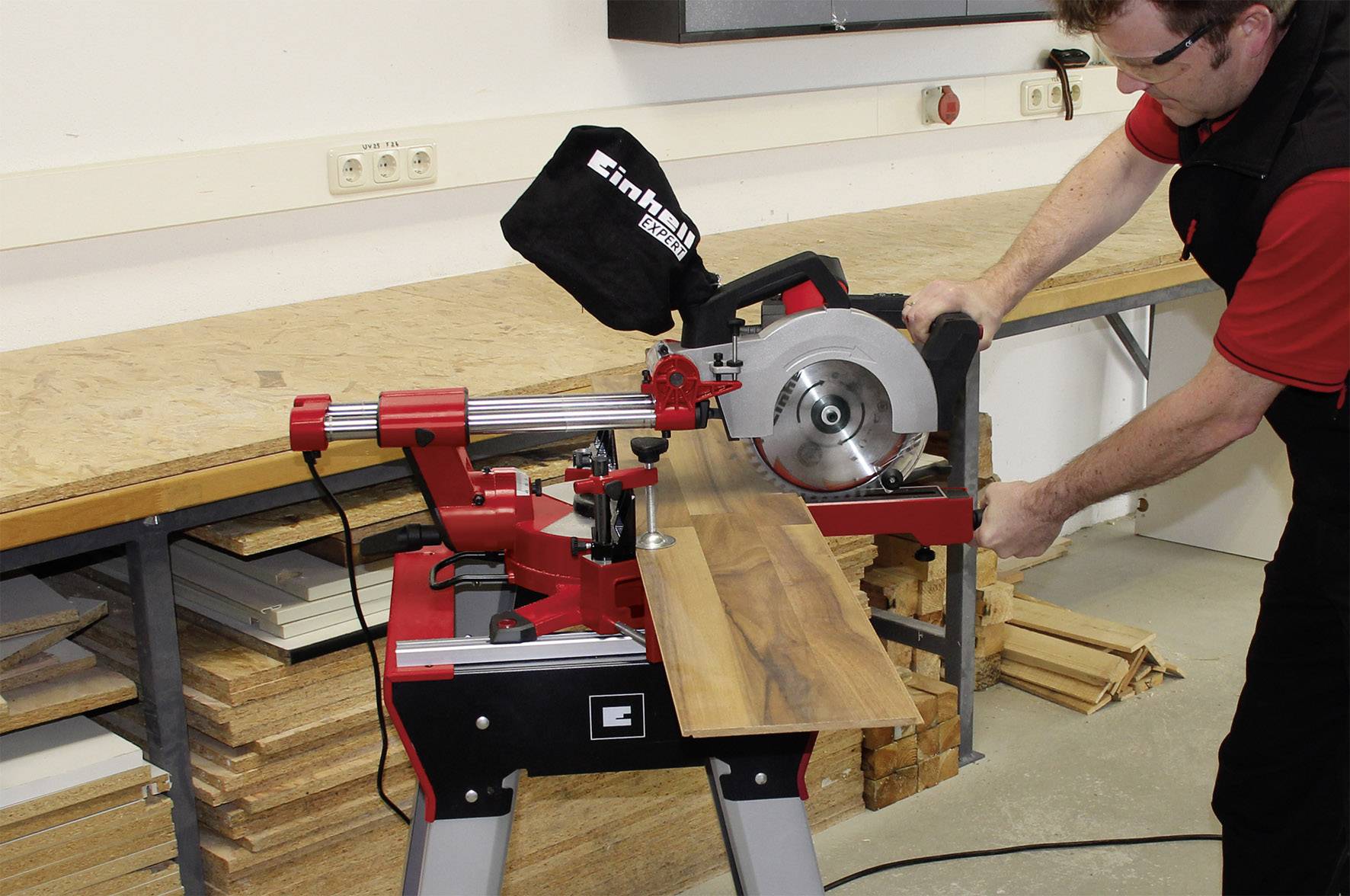 Een persoon zaagt een houten plank met een afkortzaag in een werkplaats. De werkplek is goed georganiseerd met houten materialen op de achtergrond.