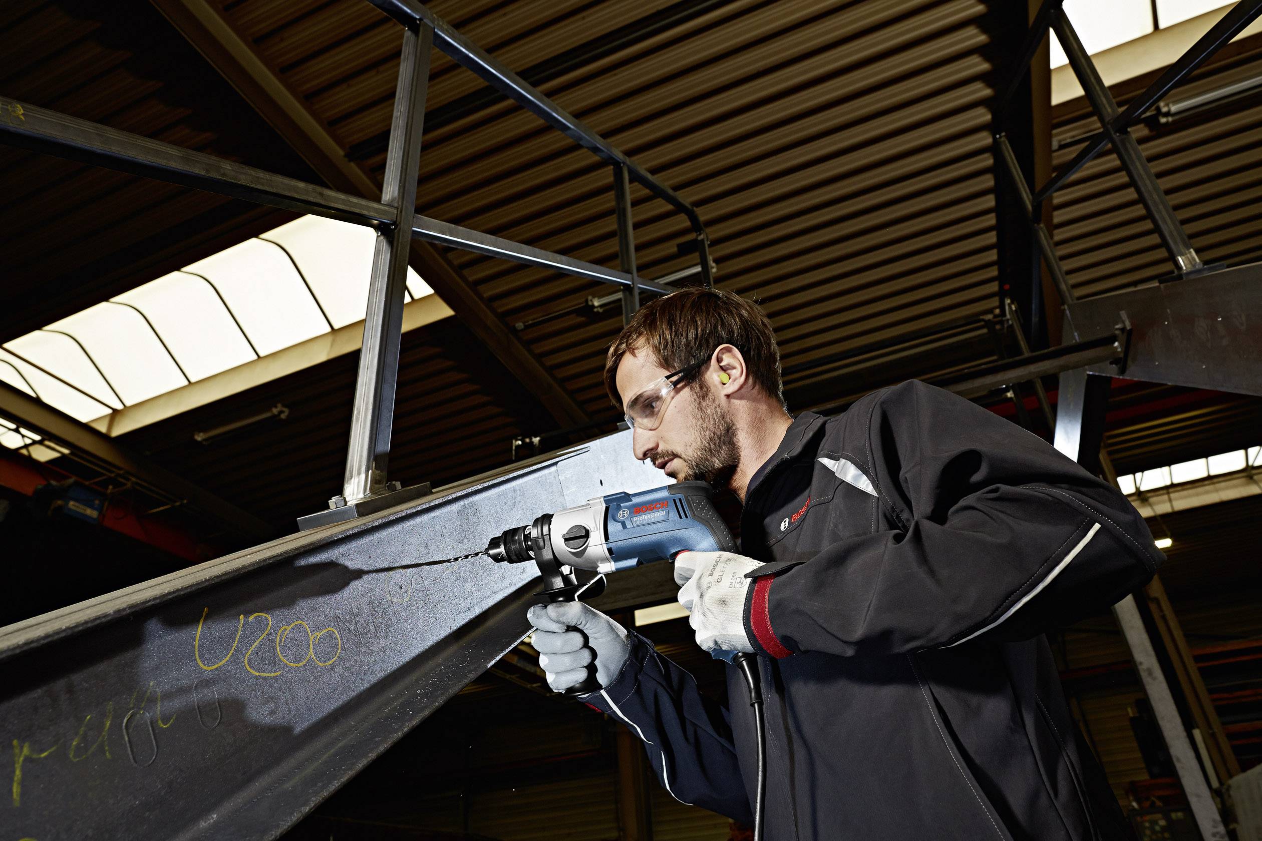 Een man boort in een stalen ligger in een industriële omgeving. Hij draagt een veiligheidsbril en handschoenen.