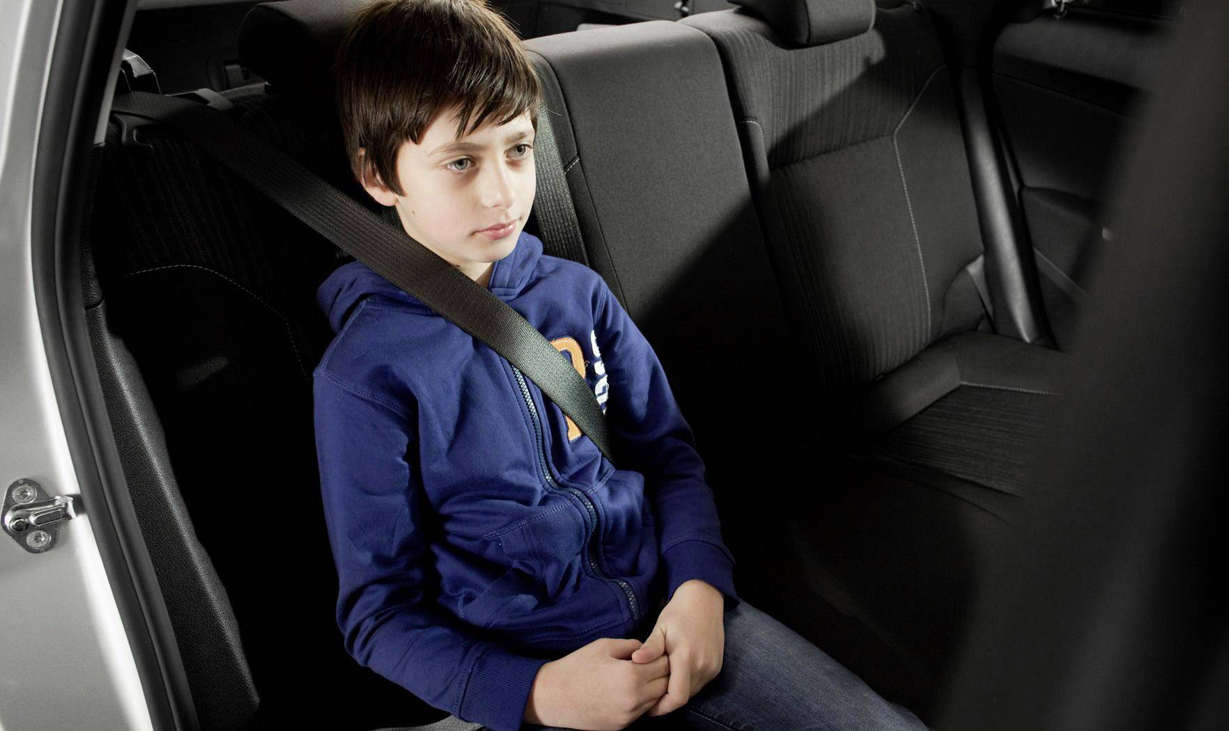 Een jongen zit op de achterbank van een auto, vastgemaakt met een veiligheidsgordel. Hij draagt een blauwe hoodie.