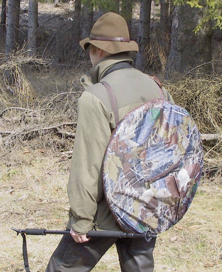 Een persoon in outdoorkleding draagt een grote, ronde gecamoufleerde tas en houdt een wandelstok vast. Op de achtergrond zijn bomen in het bos.