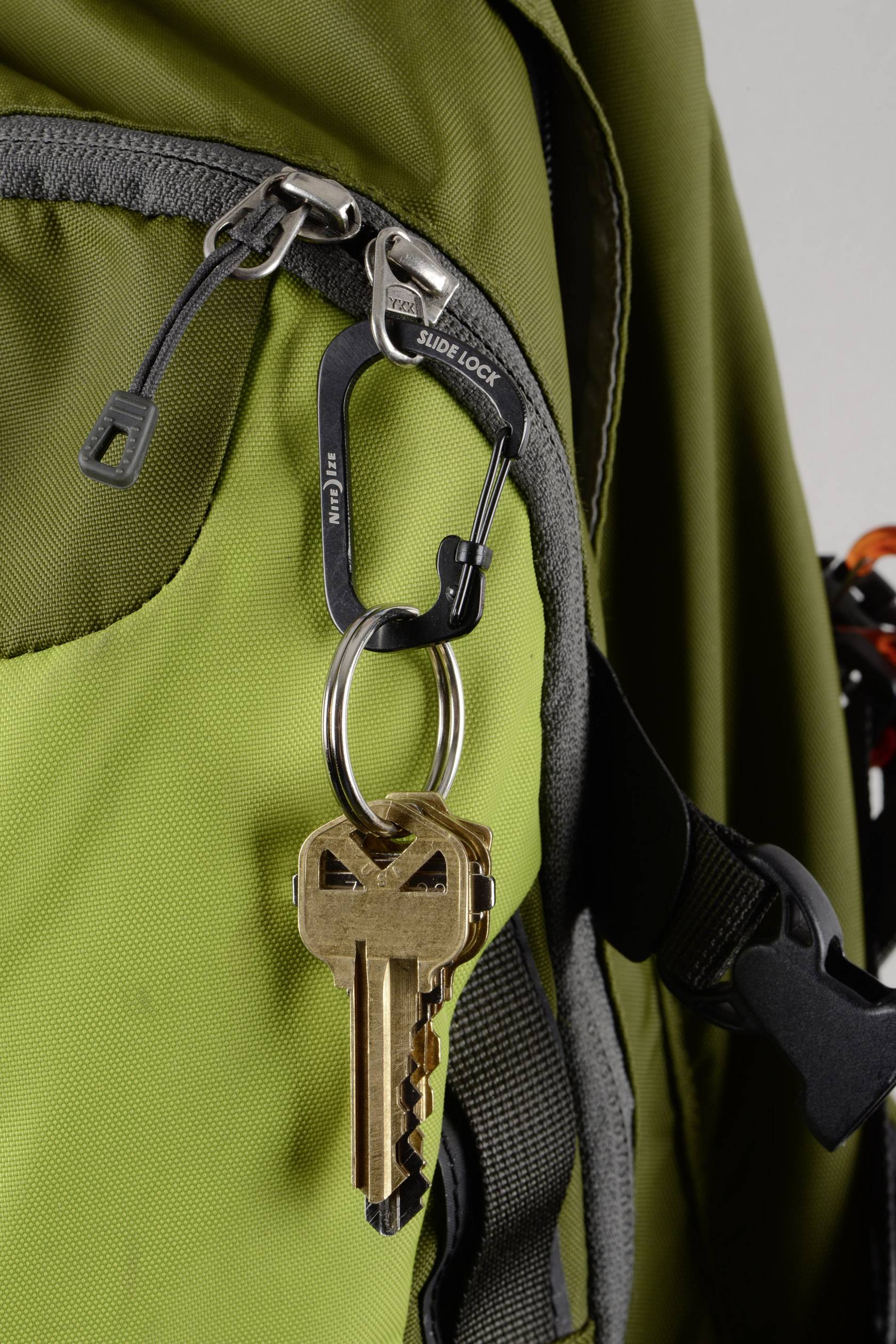 Een gouden sleutel hangt aan een zilveren karabijnhaak, die bevestigd is aan de rits van een groene rugzak.