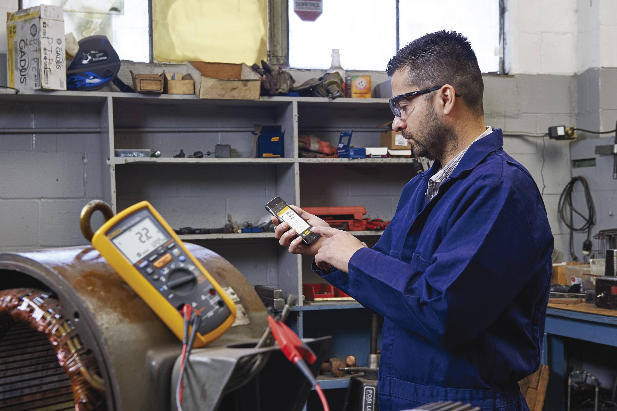 Een man in blauwe werkkleding meet de elektronica met een multimeter. Op de achtergrond zijn gereedschappen en rekken zichtbaar.