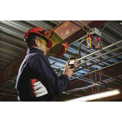 Een technicus draagt beschermende kleding en gebruikt een meetapparaat om elektrische leidingen te inspecteren in een industriële omgeving.