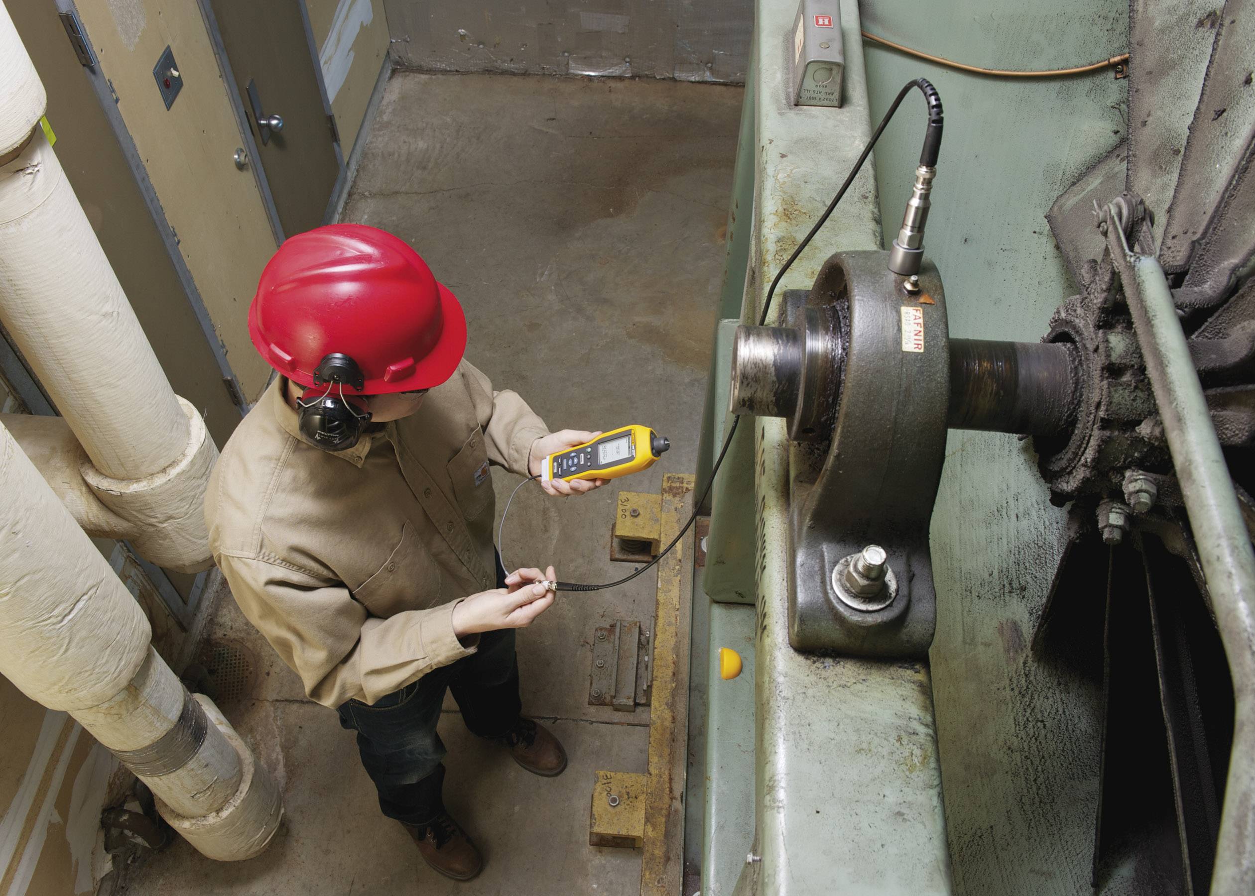 Een technicus met een rode veiligheidshelm meet de trillingen van een industriële machine met een meetapparaat om de toestand ervan te controleren.