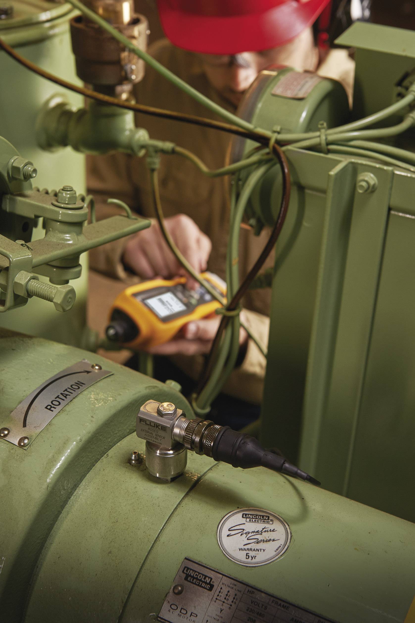 Een persoon met een rode helm controleert met een meetapparaat de staat van een groene industriemachine. Kabels zijn zichtbaar aangesloten.