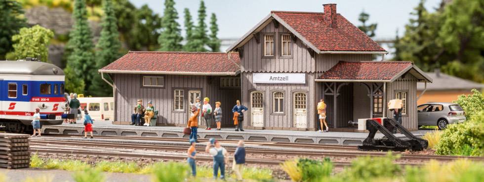 Een miniatuurstation met modelfiguren toont mensen die op de trein wachten. Een trein is te zien op het spoor, omgeven door bomen.