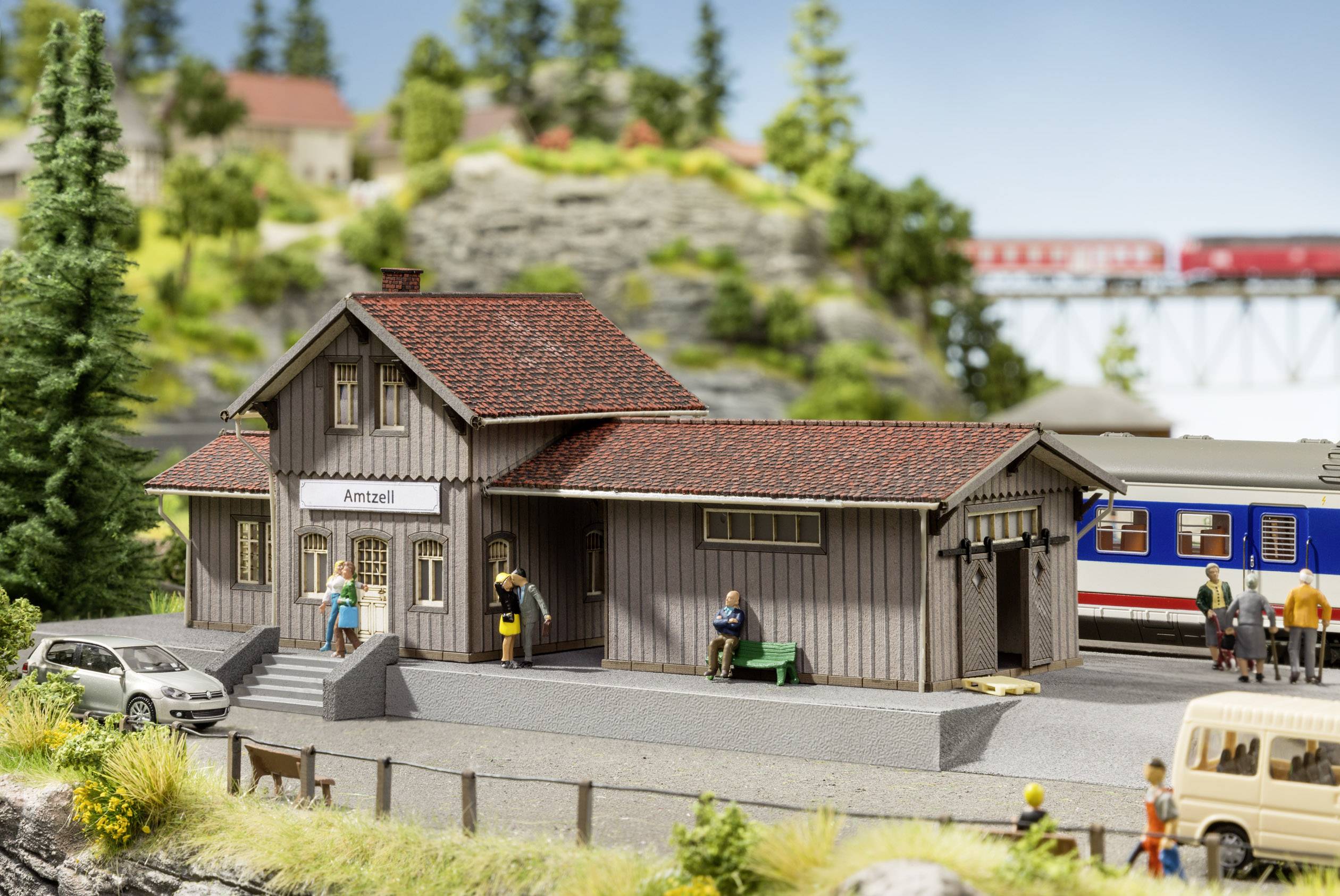 Station in een modelspoorbaan-diorama met wachtende personen en een trein op de achtergrond op een brug. Landschap met bomen en heuvels.