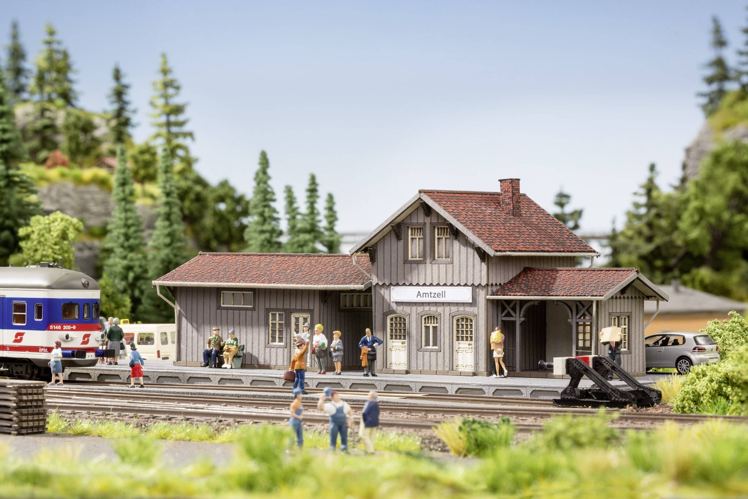 Een modelspoorbaan scène toont een klein station met wachtende modelfiguren, naaldbomen op de achtergrond en een trein langs het spoor.