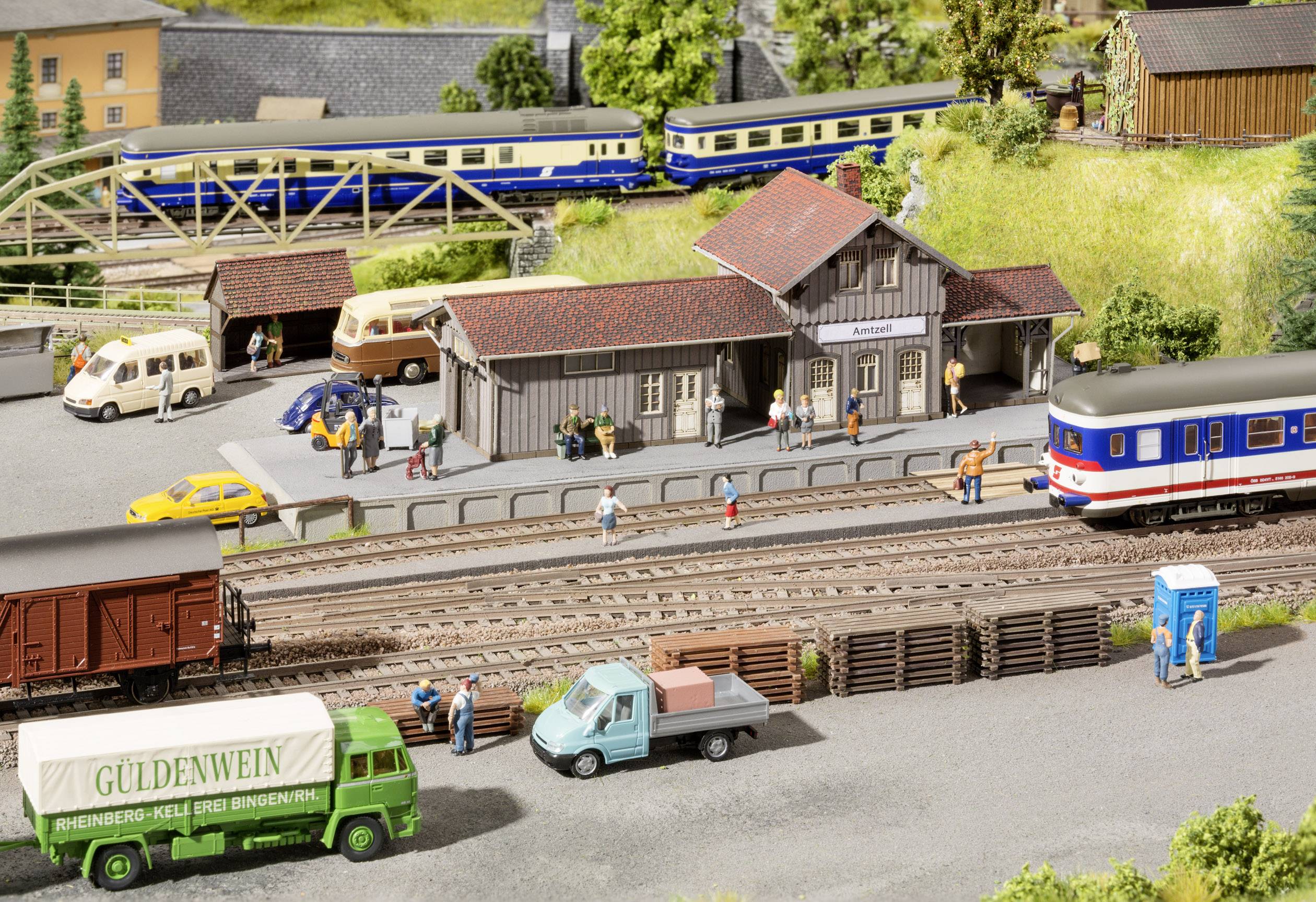 Modelspoorbaan scène met een station, treinen op de sporen, personen op het perron, een bus, meerdere auto's en een vrachtwagen op de voorgrond.