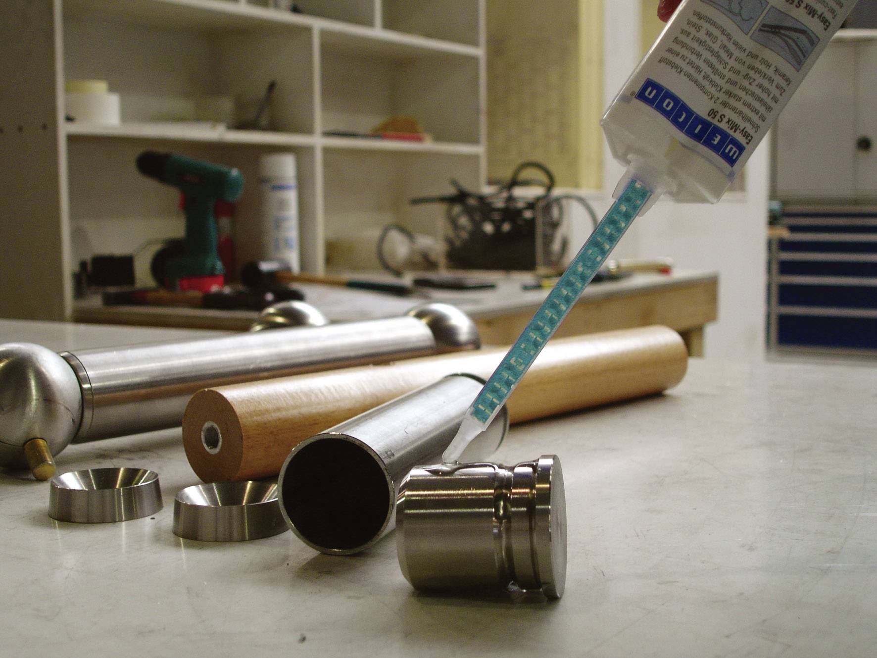 Een werkplaats met verschillende gereedschappen en materialen. Op de voorgrond zijn metalen onderdelen en een lijmtube zichtbaar.