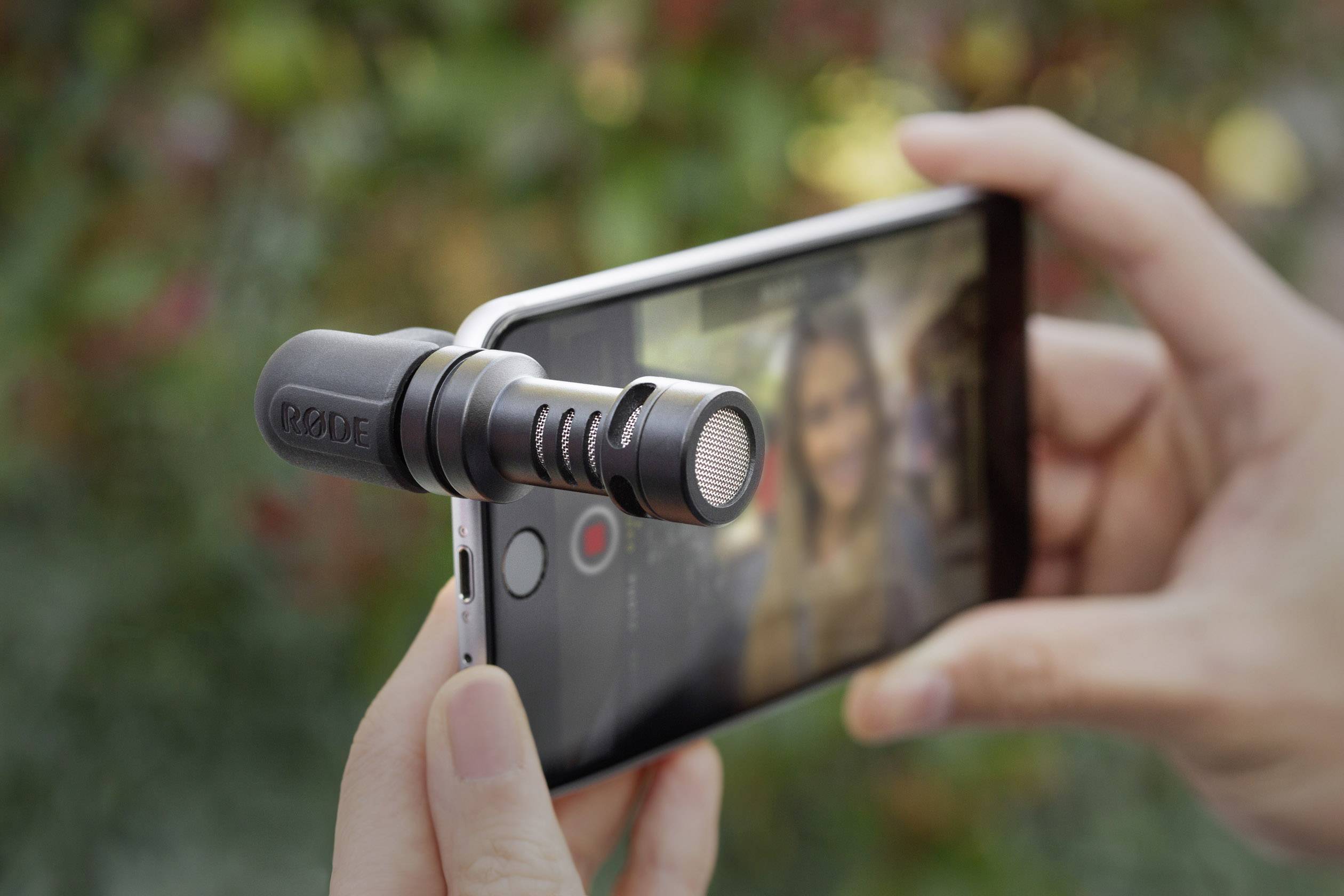 Iemand houdt een smartphone met een microfoonaccessoire, klaar voor video-opnames in de buitenlucht. Op de achtergrond is groen zichtbaar.