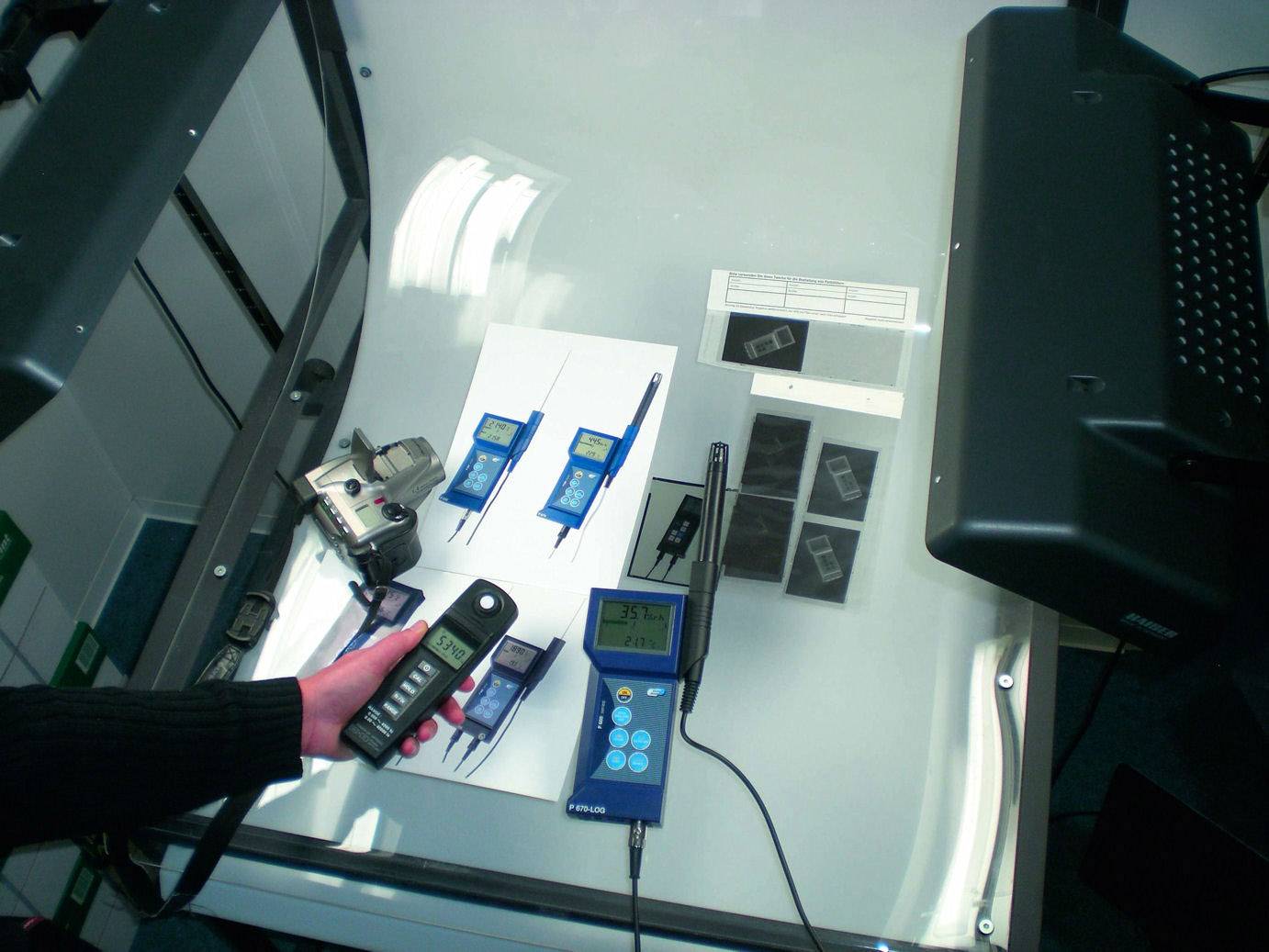 Een persoon houdt een handmeetapparaat boven een tafel met verschillende technische apparaten en documenten. Er wordt een meting uitgevoerd.
