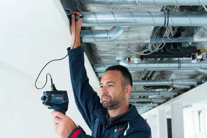 Een persoon controleert een airconditioning-unit aan het plafond met een handapparaat. De omgeving ziet er industrieel of in aanbouw uit.